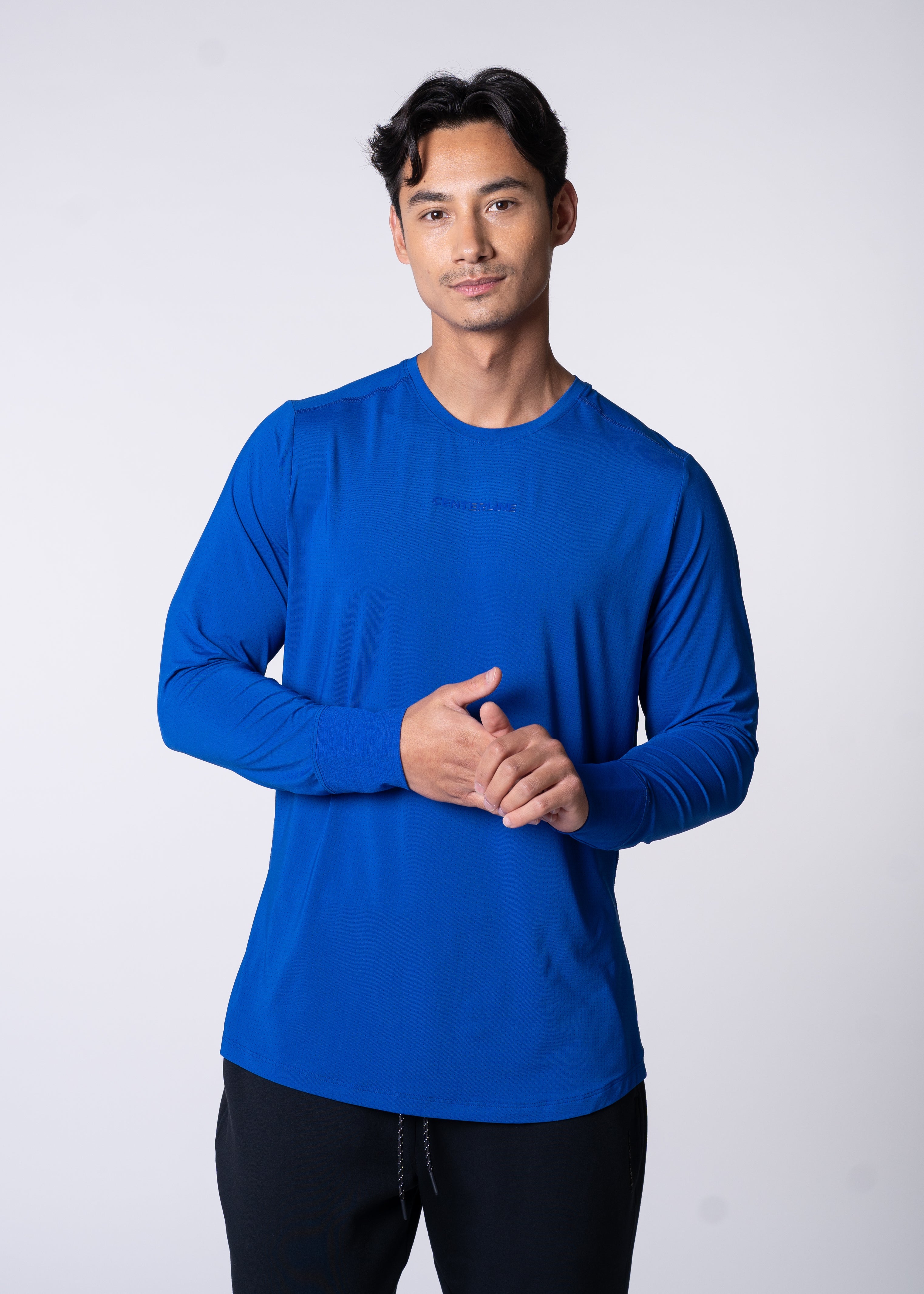 A man wearing a blue long-sleeve shirt and black pants stands against a white background.