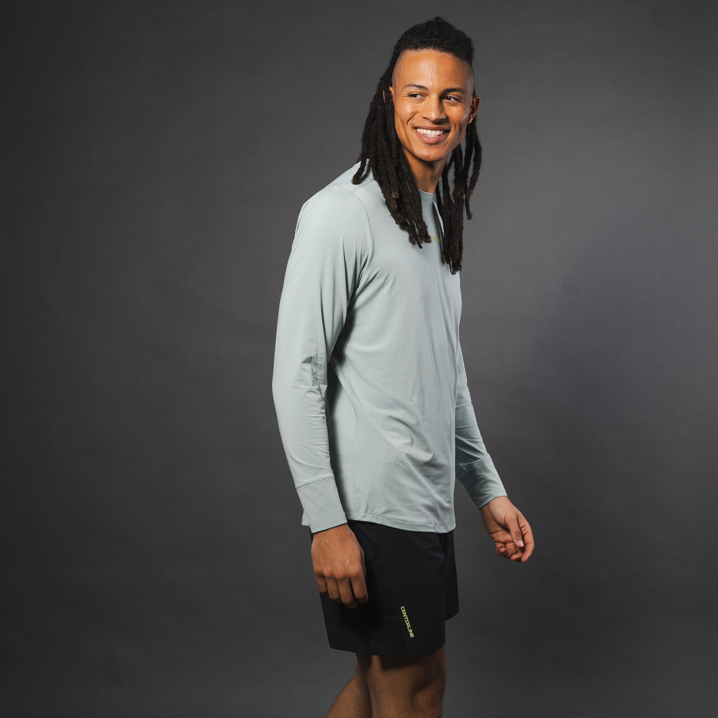 Man in activewear smiles over shoulder. Long-sleeved gray shirt and black shorts. Dreadlocks. Gray background.