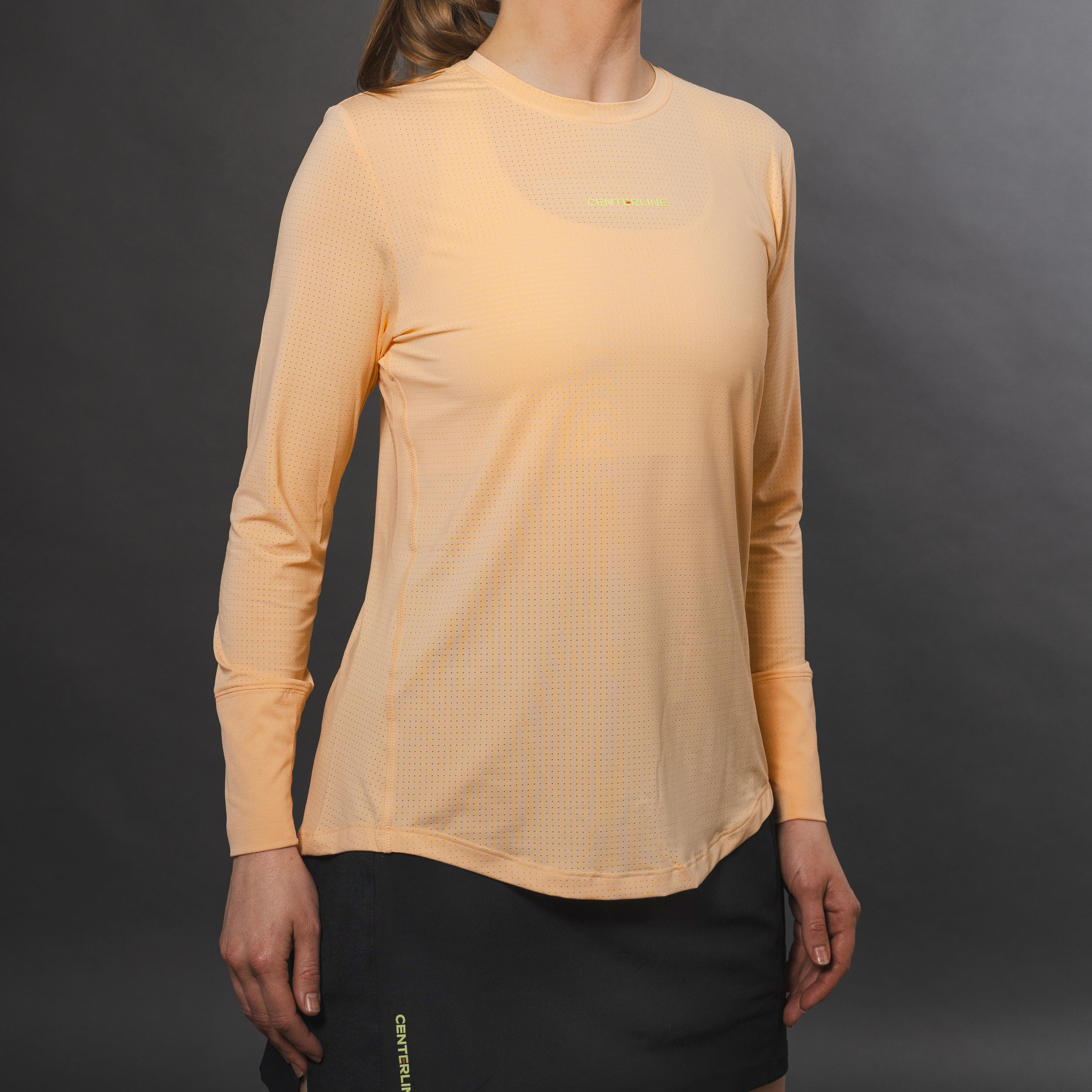 A woman wearing a long-sleeved orange athletic shirt with small perforations, and a black athletic skirt, standing against a dark gray background.