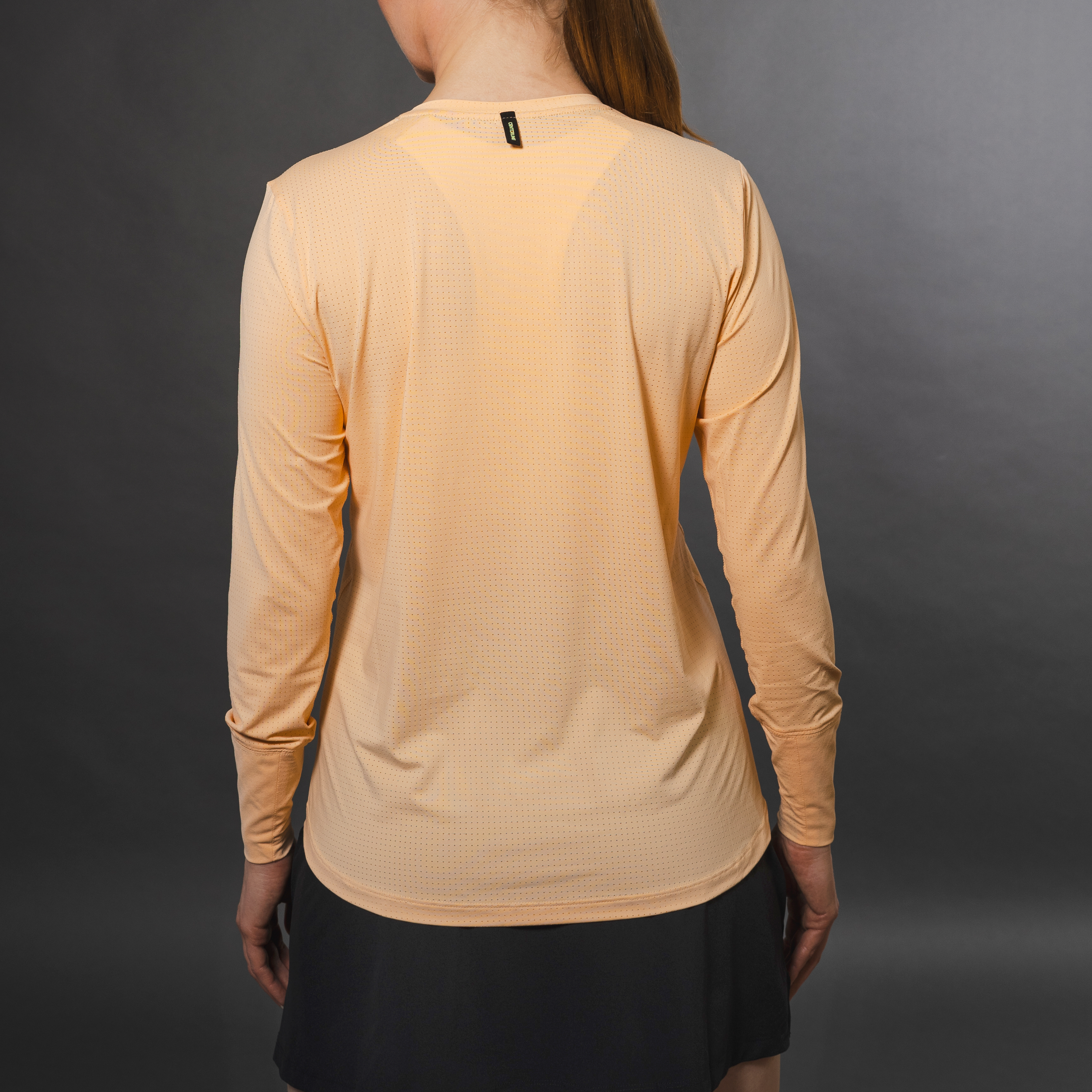 A woman in a peach long-sleeved shirt and a dark skirt stands against a grey background.