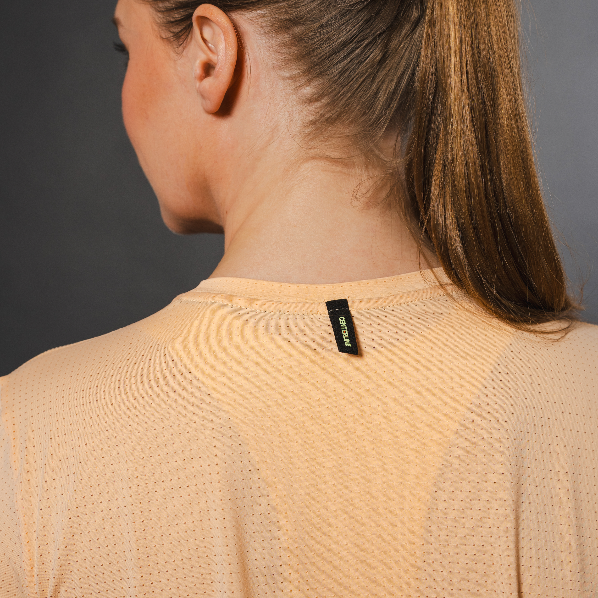 Back of a woman wearing a peach-colored, perforated athletic shirt with a black brand tag. The woman has blonde hair in a ponytail and is facing away from the camera.