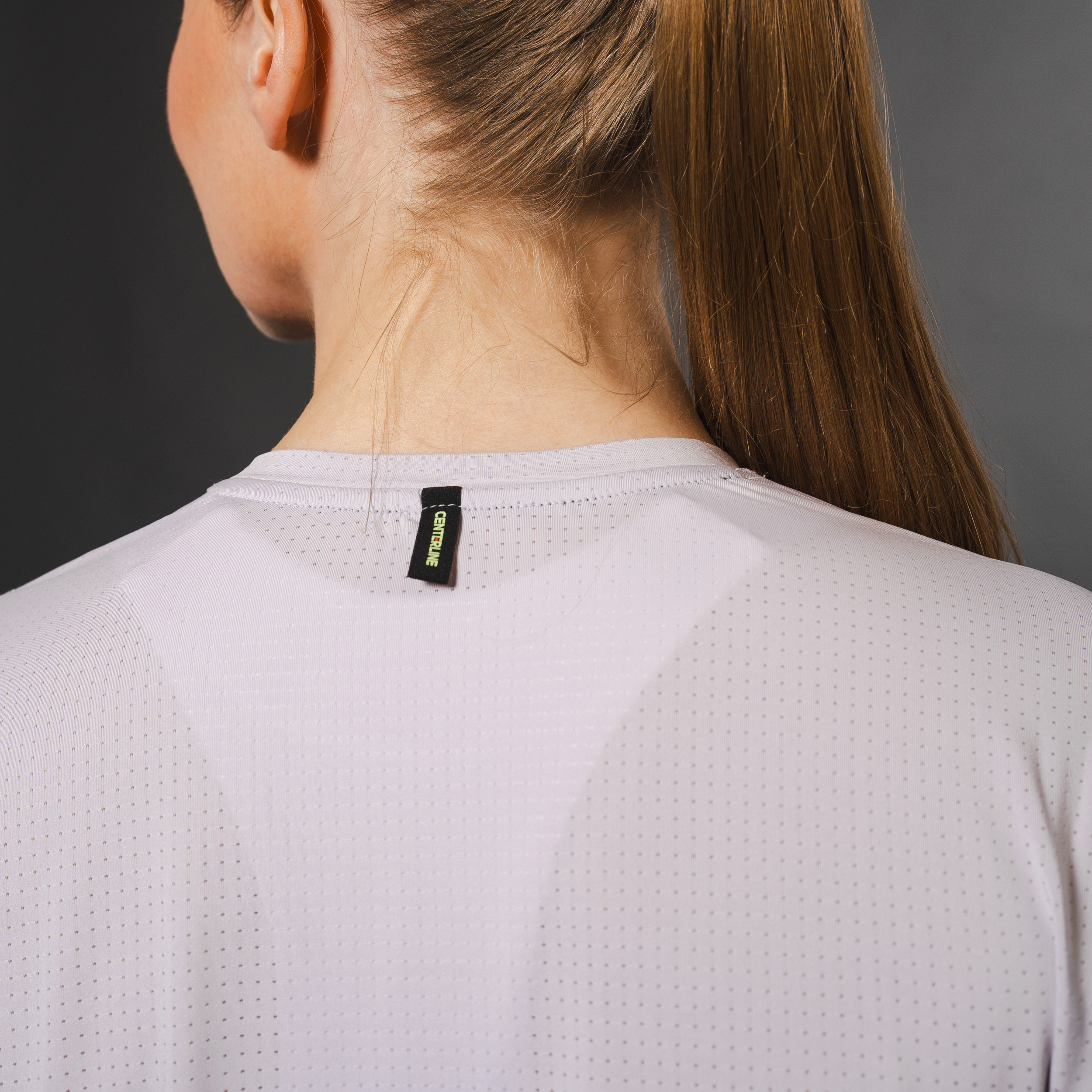 Woman's back in a grey, perforated athletic shirt, with a black tag on the neck.