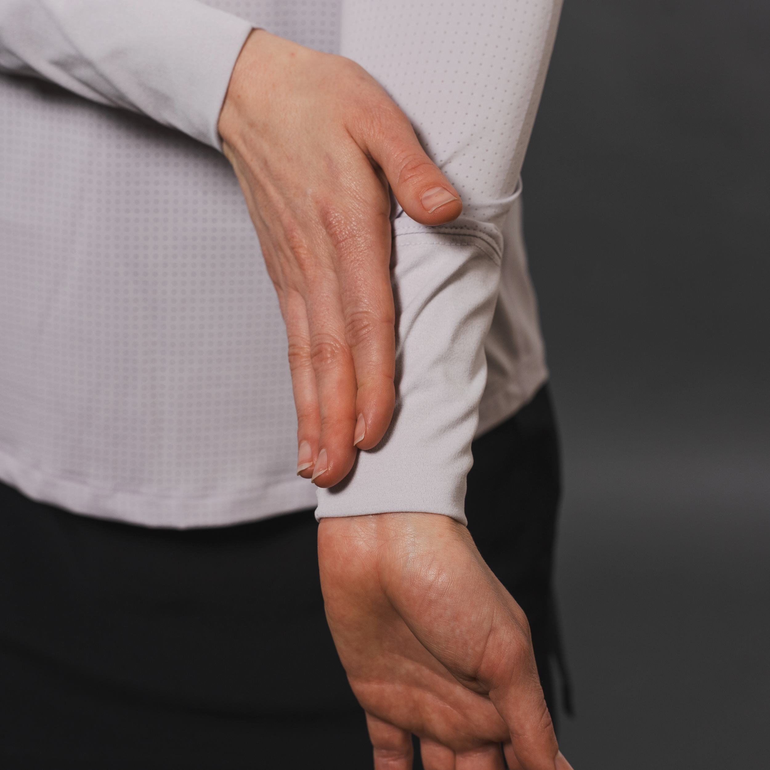 Person wearing a pale purple long-sleeved shirt with a textured pattern. The person's arm is bent with one hand resting on the wrist of the other arm. They are wearing a dark skirt. The background is gray.