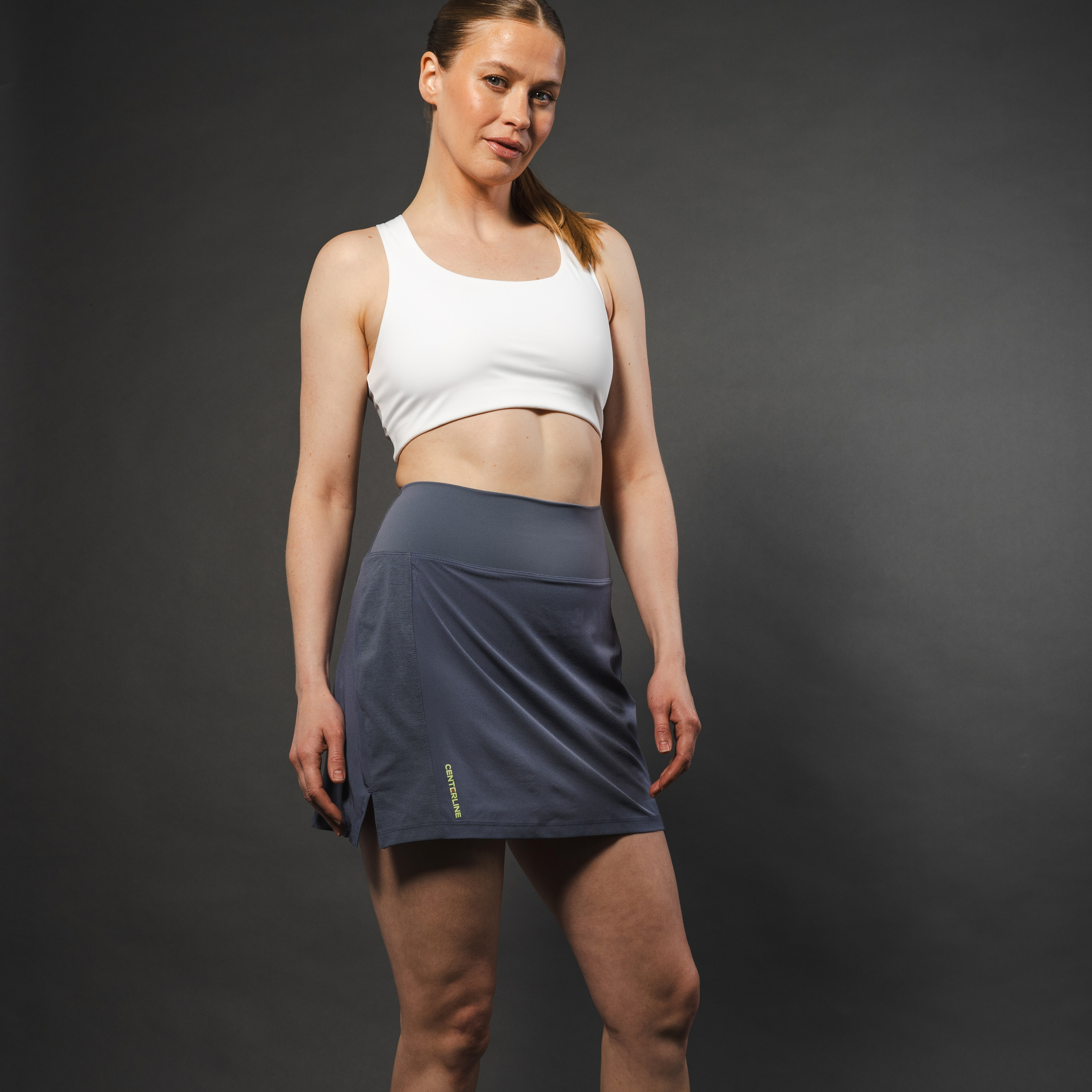 Woman in white sports bra and gray skort against gray background.