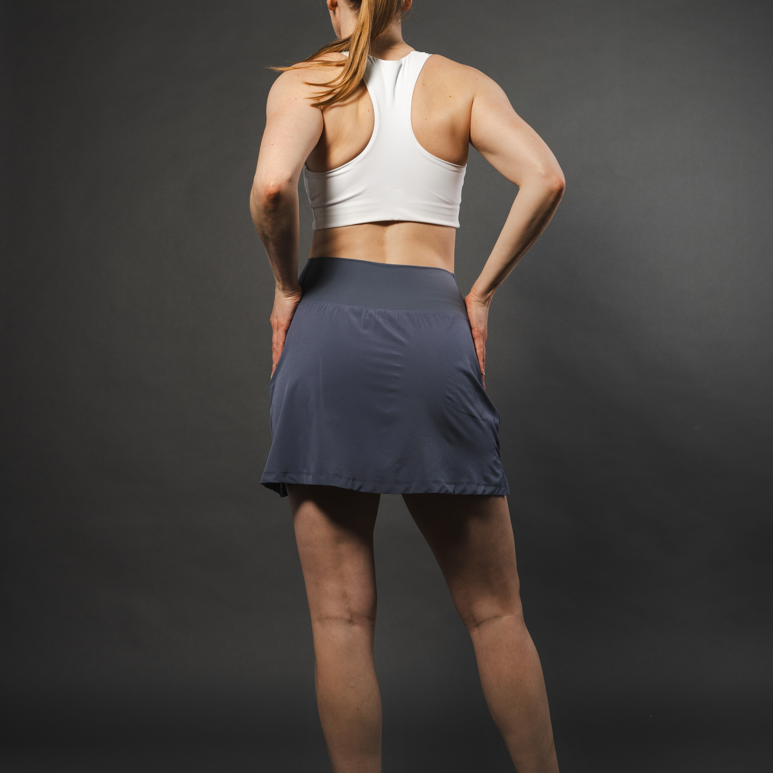 Woman in white sports bra and gray skirt, hands on hips, rear view, gray background.