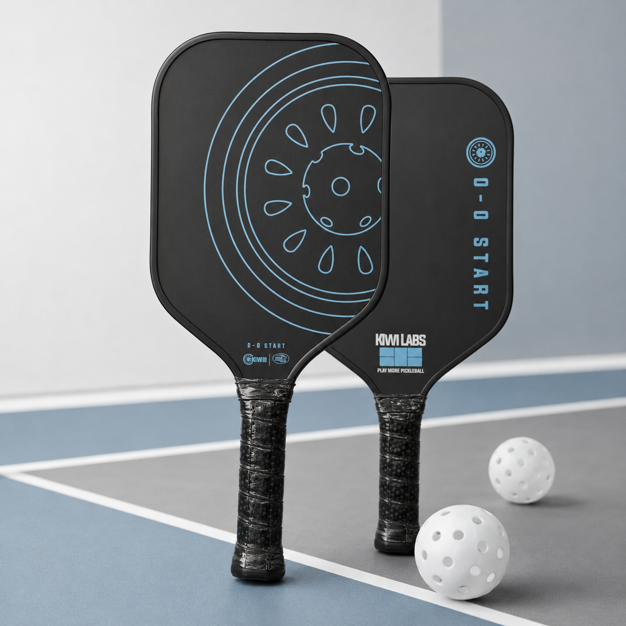 Two black pickleball paddles by KiwiLabs Pickleball, with blue graphics and textured grips, standing on a court with two white pickleballs.