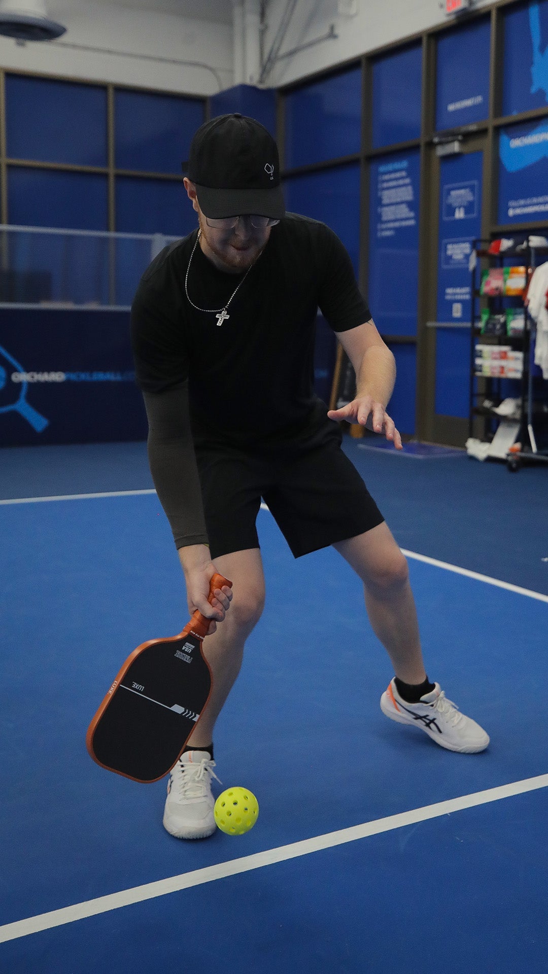 A man in black athletic wear and a hat plays pickleball on a blue court.