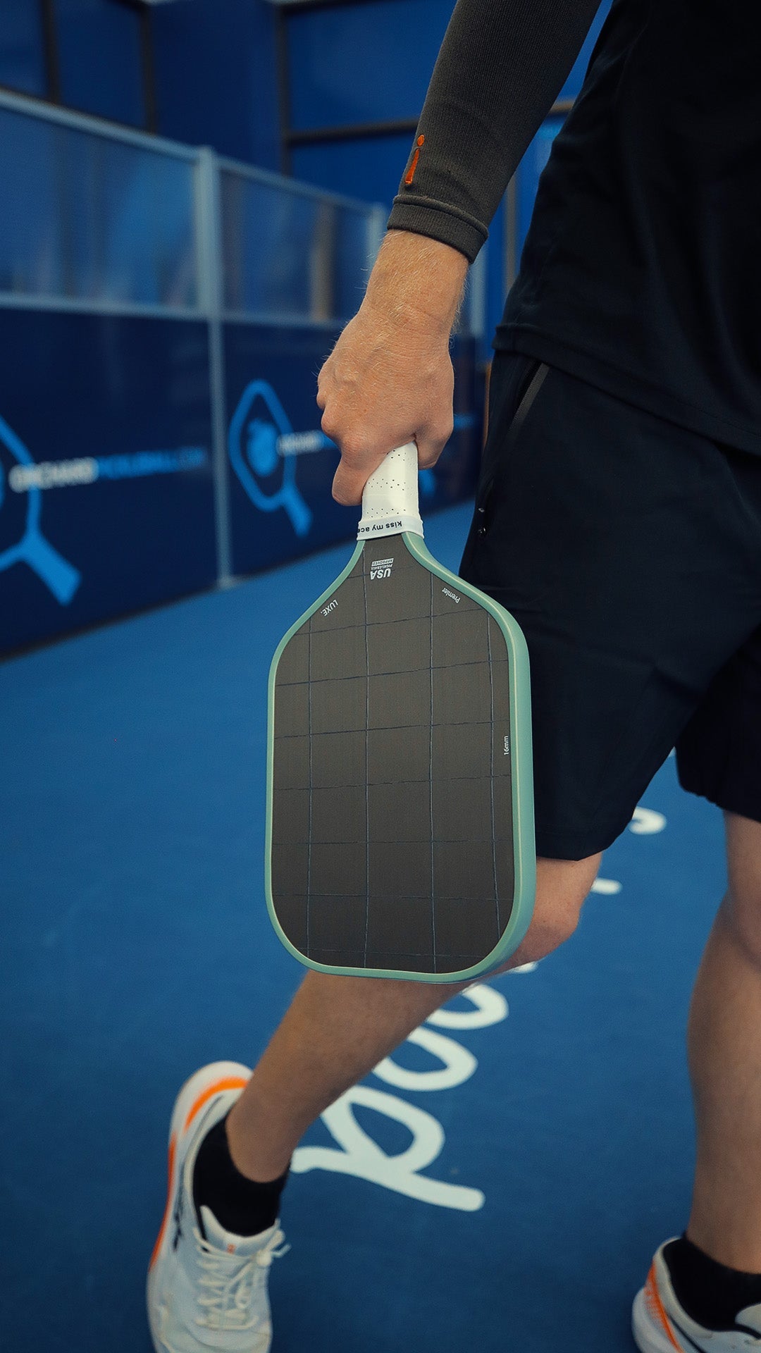 A person holding a pickleball paddle, wearing black athletic shorts, a gray athletic sleeve, and white and orange sneakers, standing on a blue surface with white lettering.