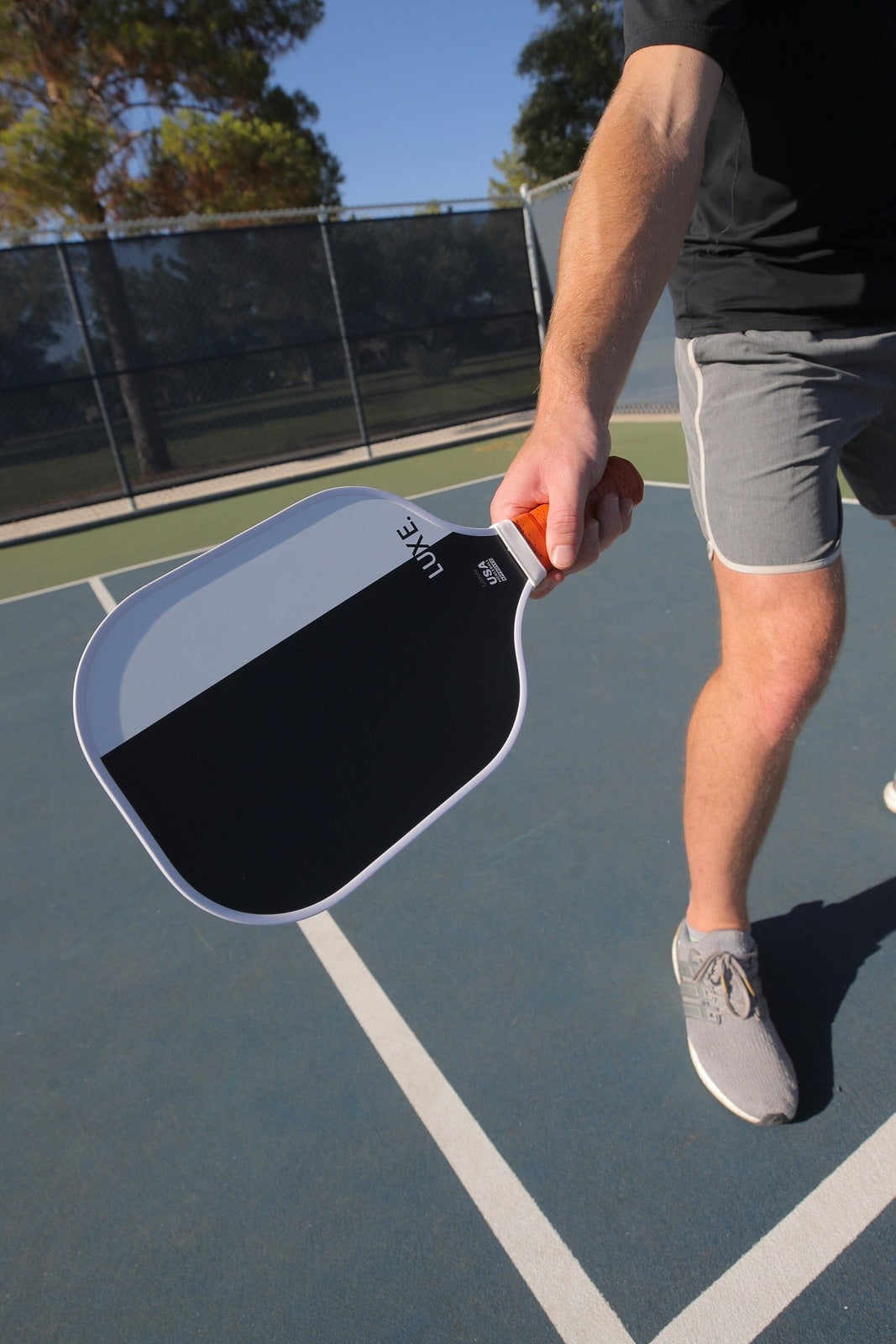 A man holding a black and grey Luxe pickleball paddle on a blue tennis court. He is wearing grey shorts and grey tennis shoes.