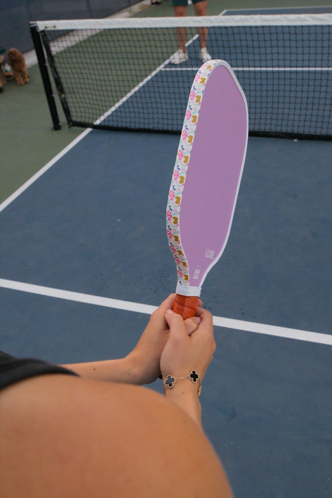 A person holding a purple pickleball paddle with a decorated edge on a blue and green court. A tennis net is in the background. The person is wearing a clover-shaped bracelet.