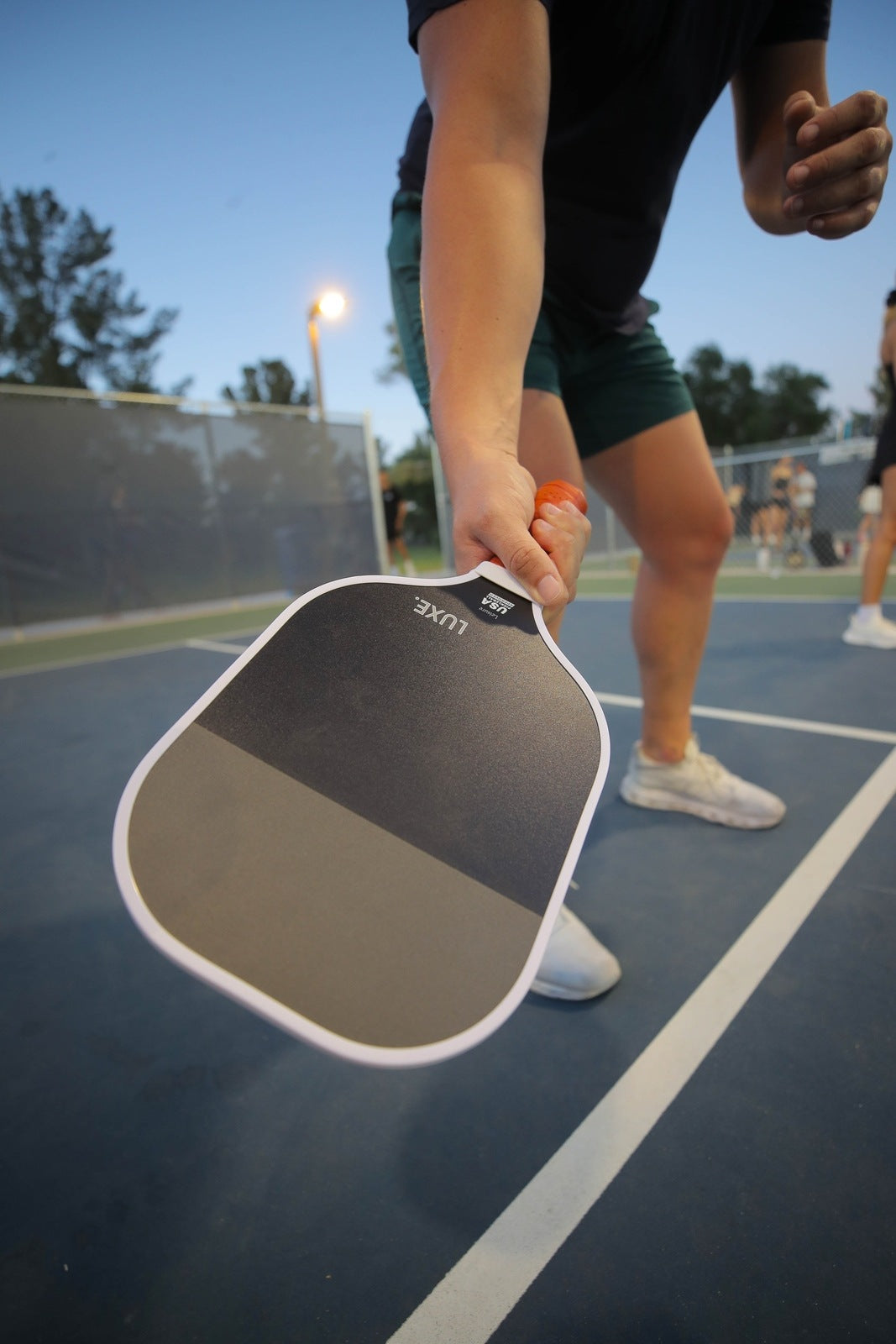 Person holding a black and gray Luxe pickleball paddle on a blue outdoor court, wearing a black shirt, green shorts, and white sneakers.
