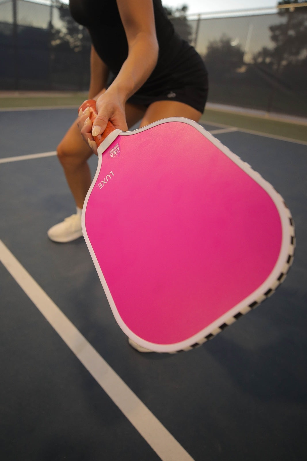 Woman in black athletic clothes holding pink pickleball paddle on a blue court.