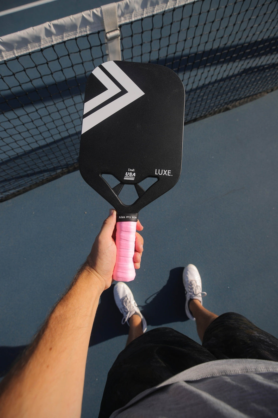 Person holding a black pickleball paddle with a pink grip, wearing white shoes, and standing on a blue court.