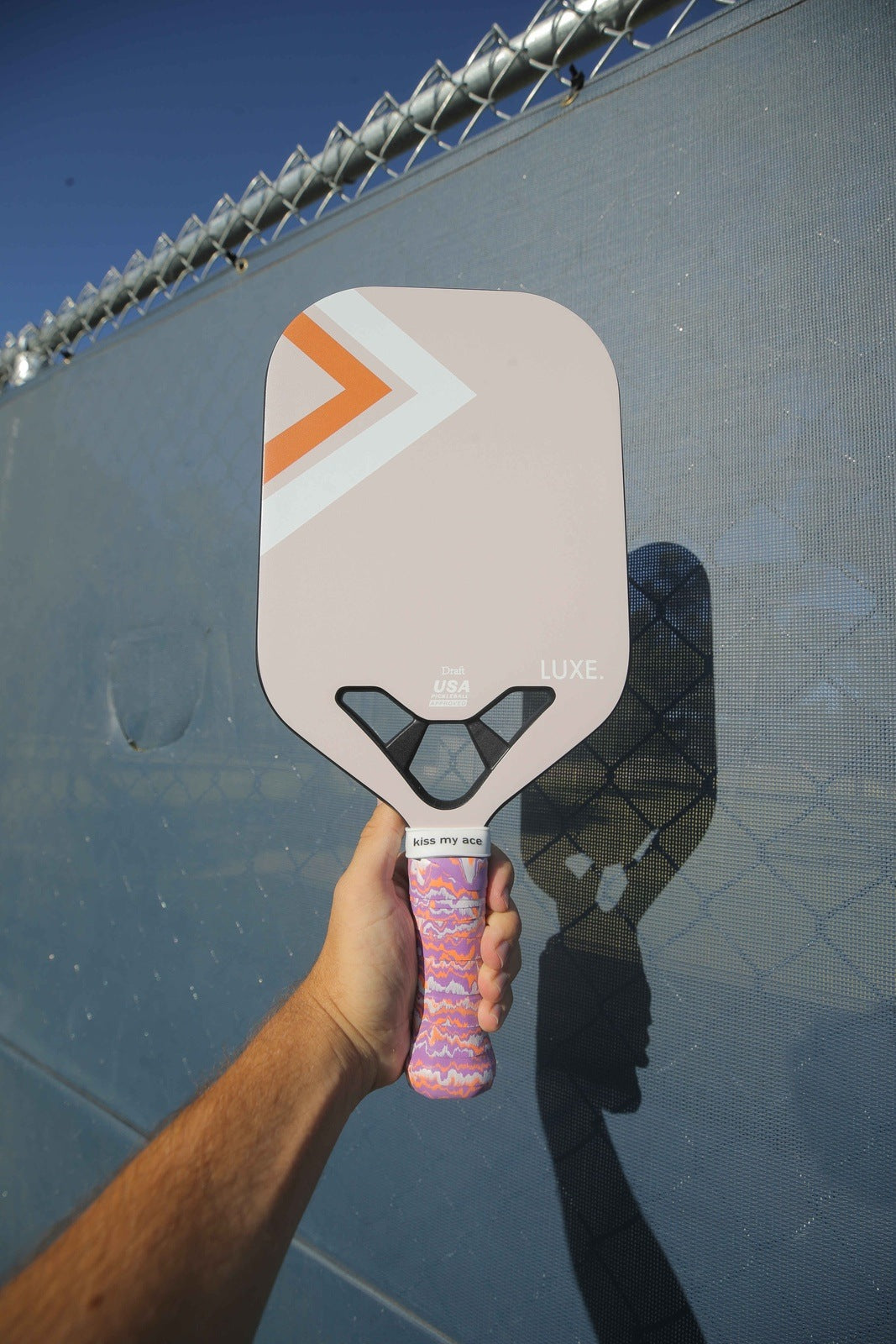 A hand holds a pink and orange pickleball paddle in front of a chain-link fence.