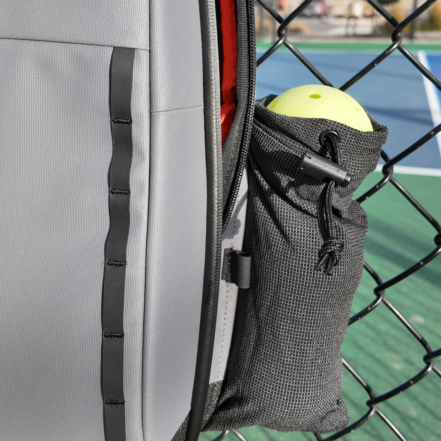 Grey textured Pickleball Sling by FORWRD, with an attached black mesh pouch holding a yellow pickleball, against a chain-link fence.