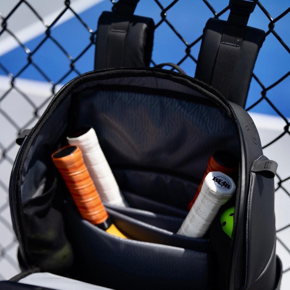 Black pickleball bag by FORWRD, open to show organized compartments holding paddles and a green pickleball against a chain-link fence.