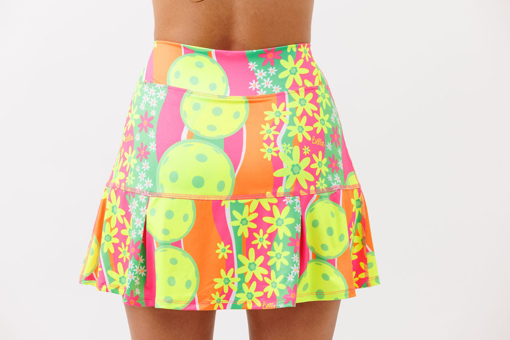 Brightly patterned pleat skort by Pickleball Bella, featuring neon yellow pickleball balls, pink and yellow flowers, and vertical stripes.