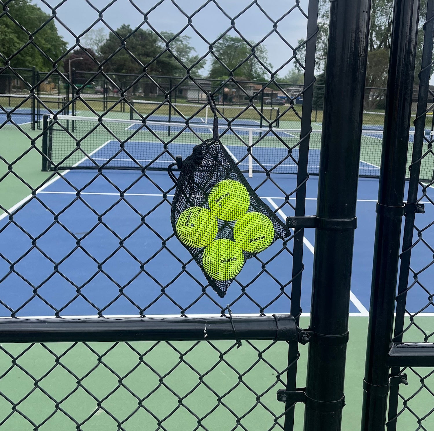 Black mesh bag of yellow pickleballs hanging on a black chain link fence in front of a green and blue court.