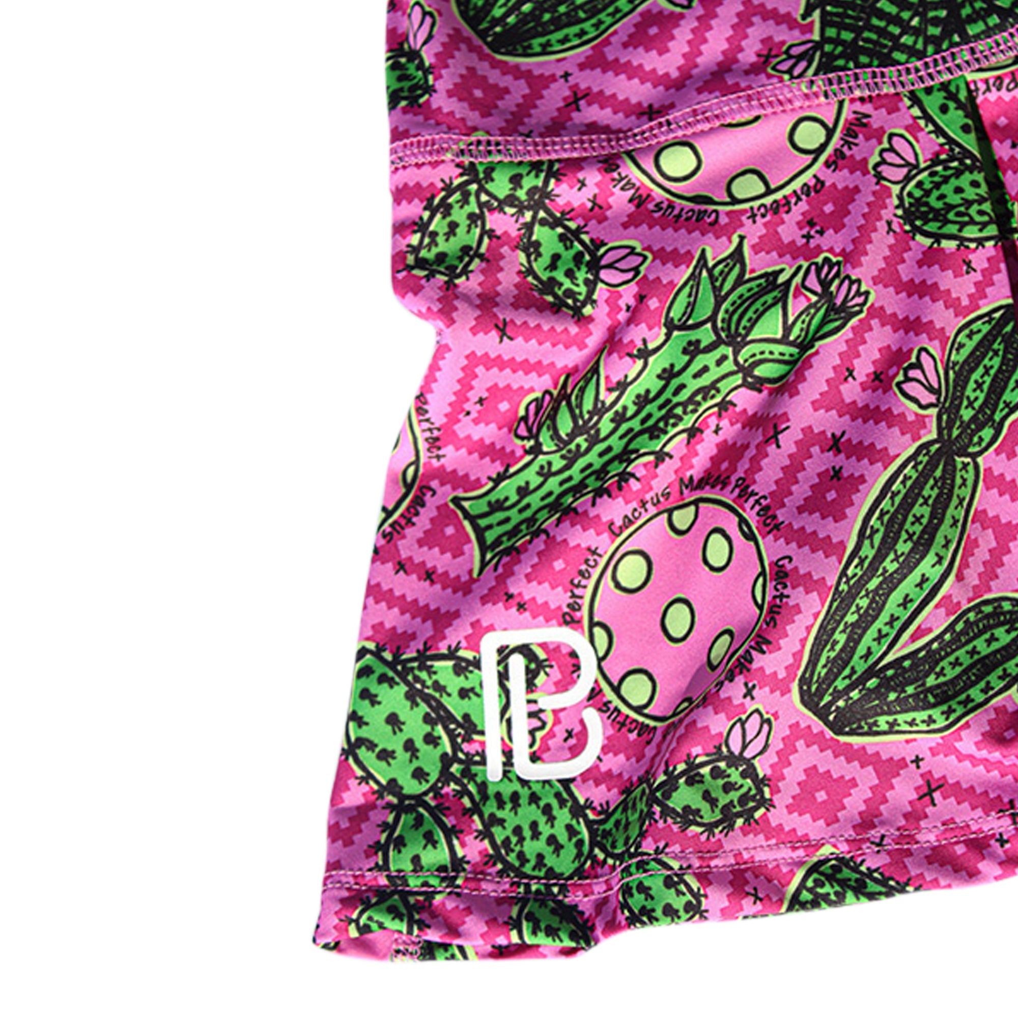 Cropped view of a pink pleat skort by Pickleball Bella, featuring a green cactus pattern, 'PB' logo, and 'Cactus Makes Perfect' text.