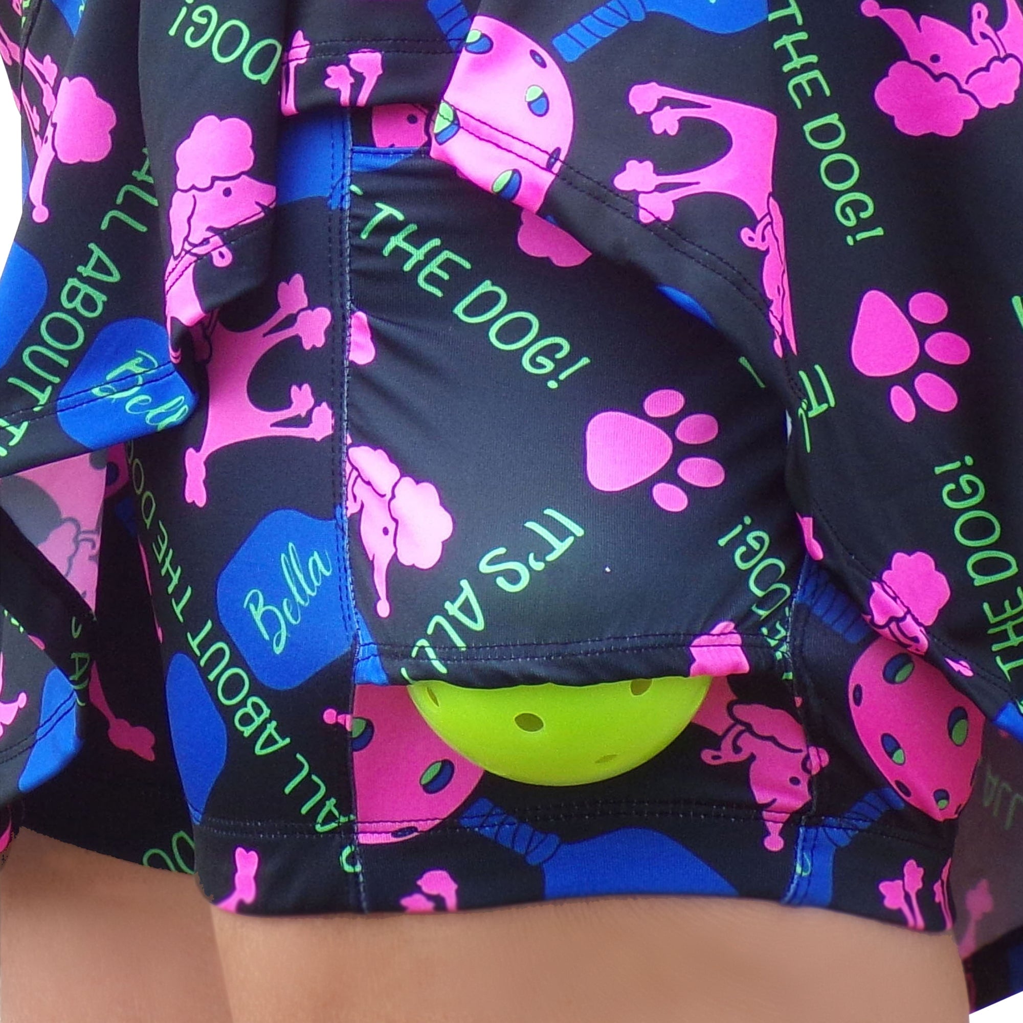 Black drop pleat skort by Pickleball Bella, patterned with pink poodles, paw prints, blue paddles, and green text, holding a yellow pickleball.
