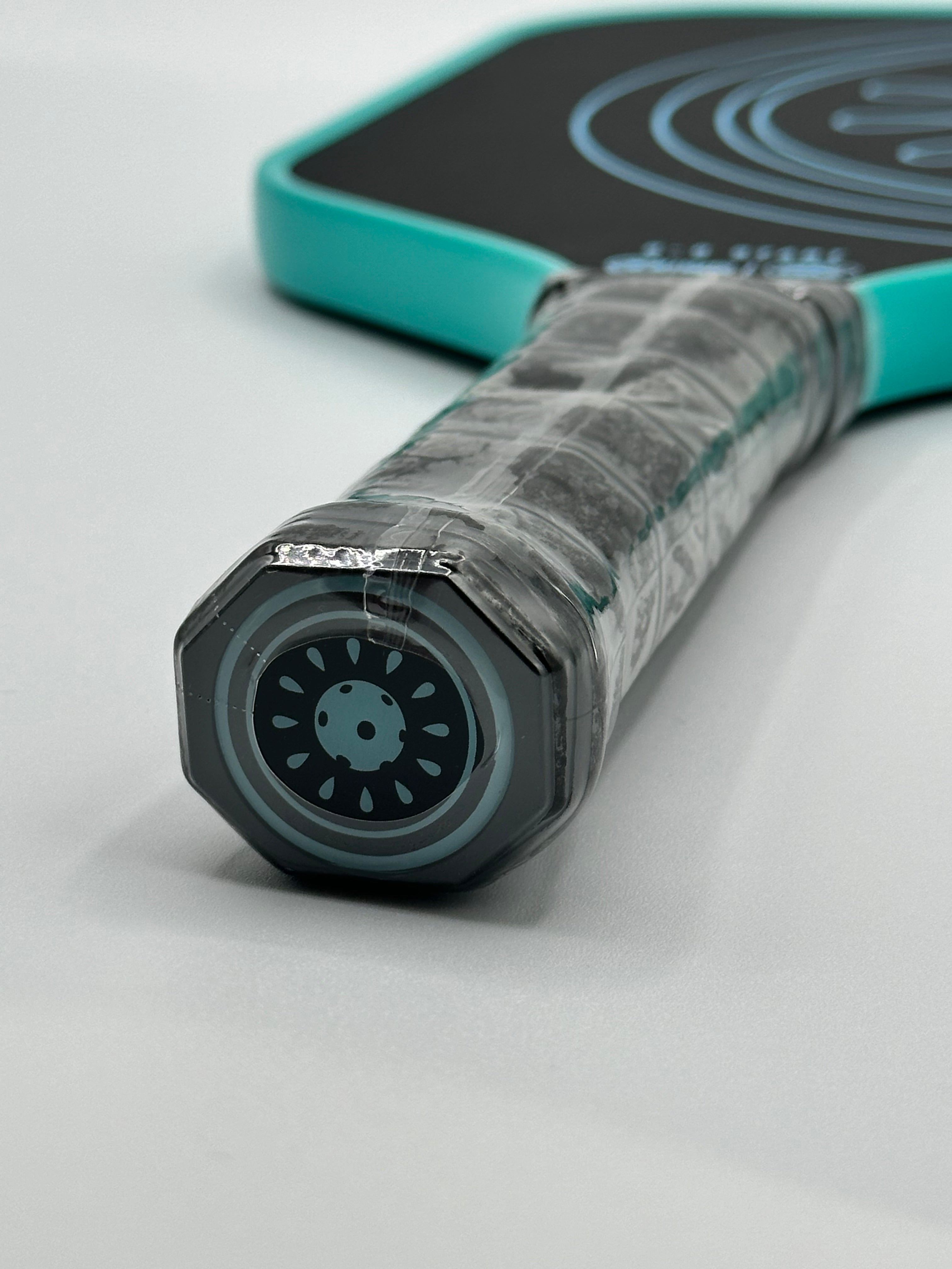 Close up of the grip and bottom of a turquoise and black pickleball paddle.