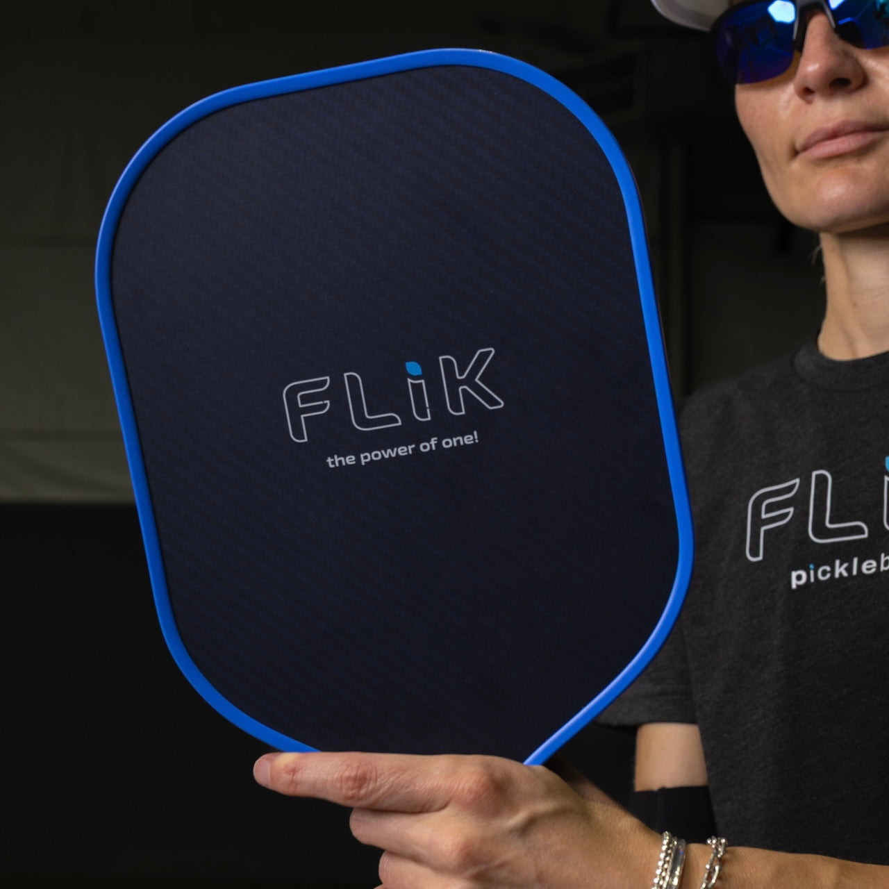 A person holding a black and blue pickleball paddle with the "Flik" logo, wearing sunglasses and a gray shirt that also features a similar logo.