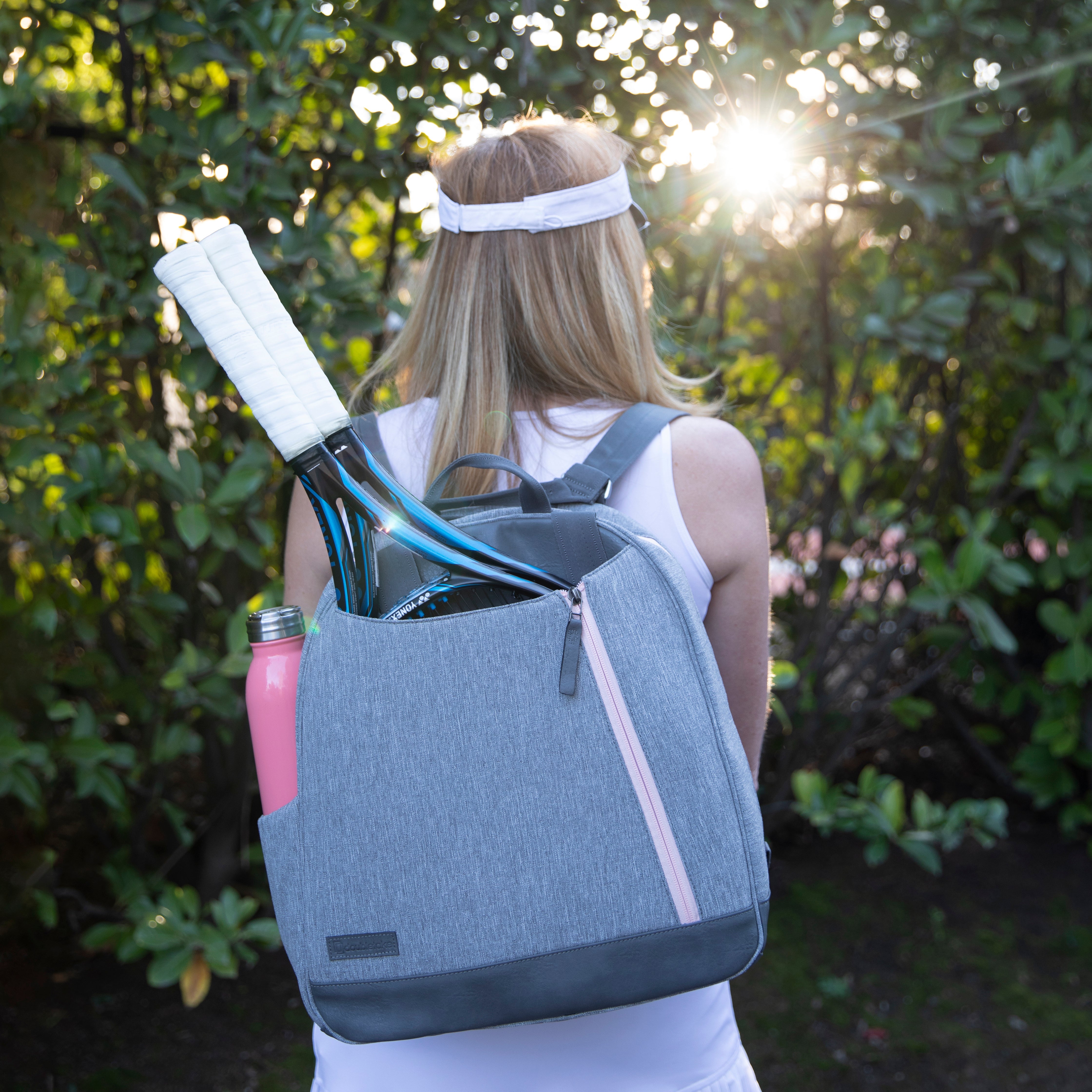 Grey tennis backpack by Doubletake, worn on a woman's back, featuring two tennis rackets, a pink water bottle, and a pink zipper.