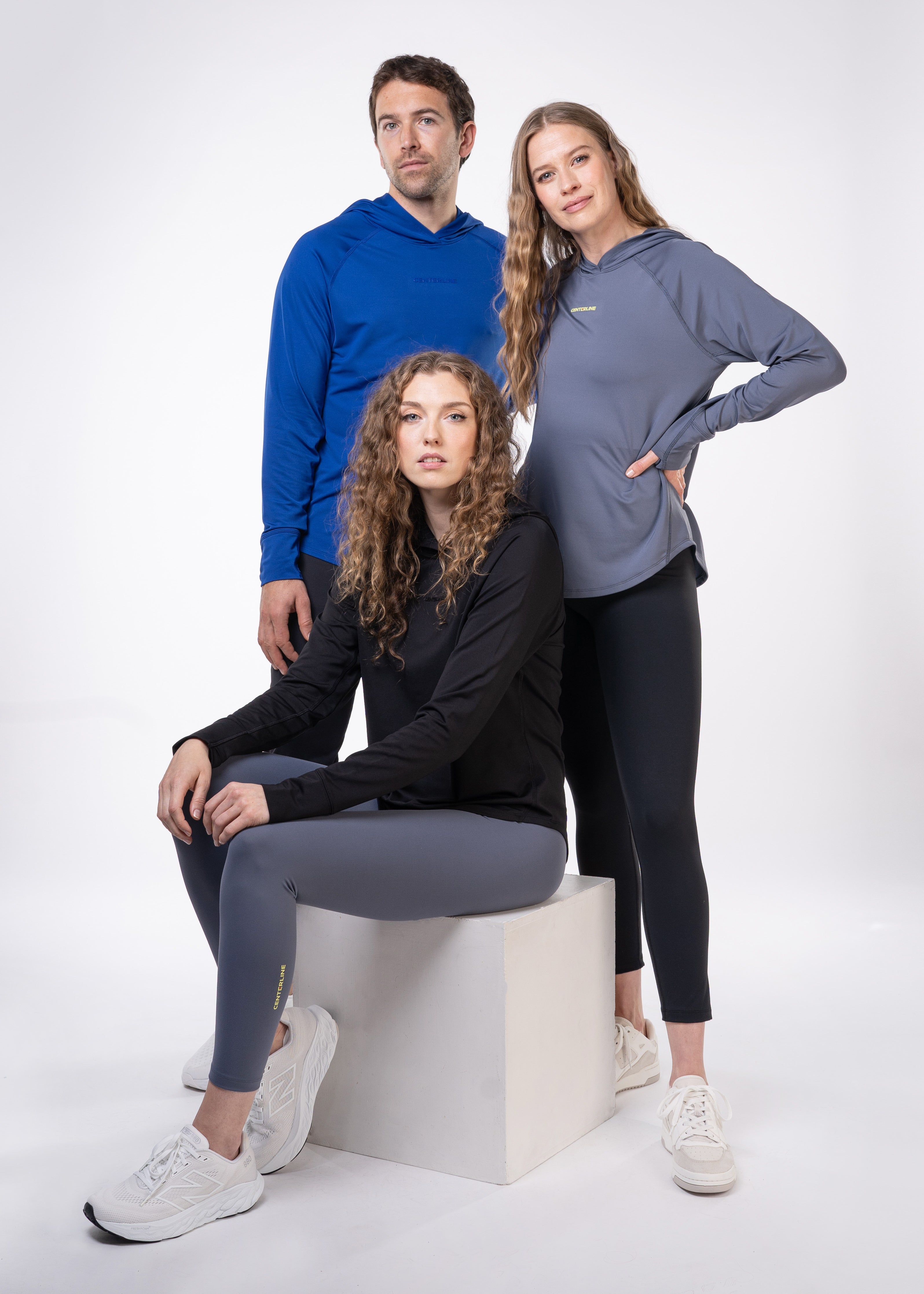 Three models wearing activewear. A woman is sitting on a white cube. A man in a blue hoodie and a woman in a grey hoodie stand behind her. Each person is wearing matching leggings and athletic shoes.