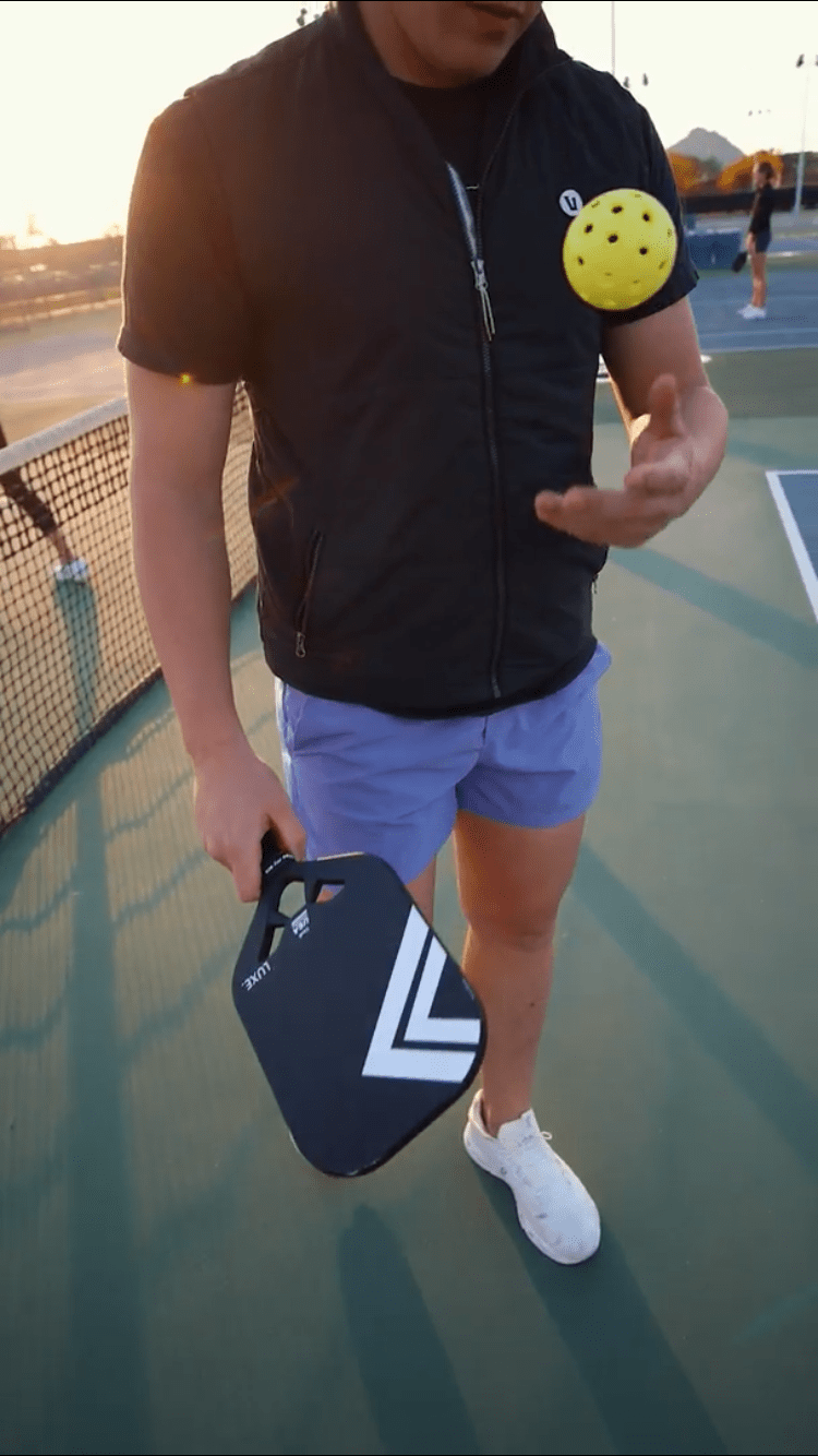 Man in vest and blue shorts juggling yellow pickleball with black paddle on court.