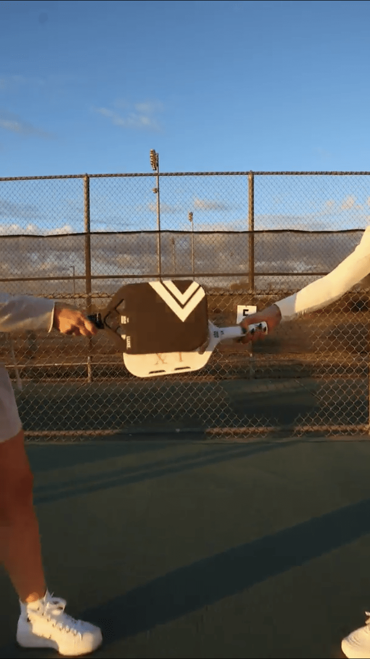 Black and white pickleball paddle with V-shaped design, held by two players on outdoor court, side view
