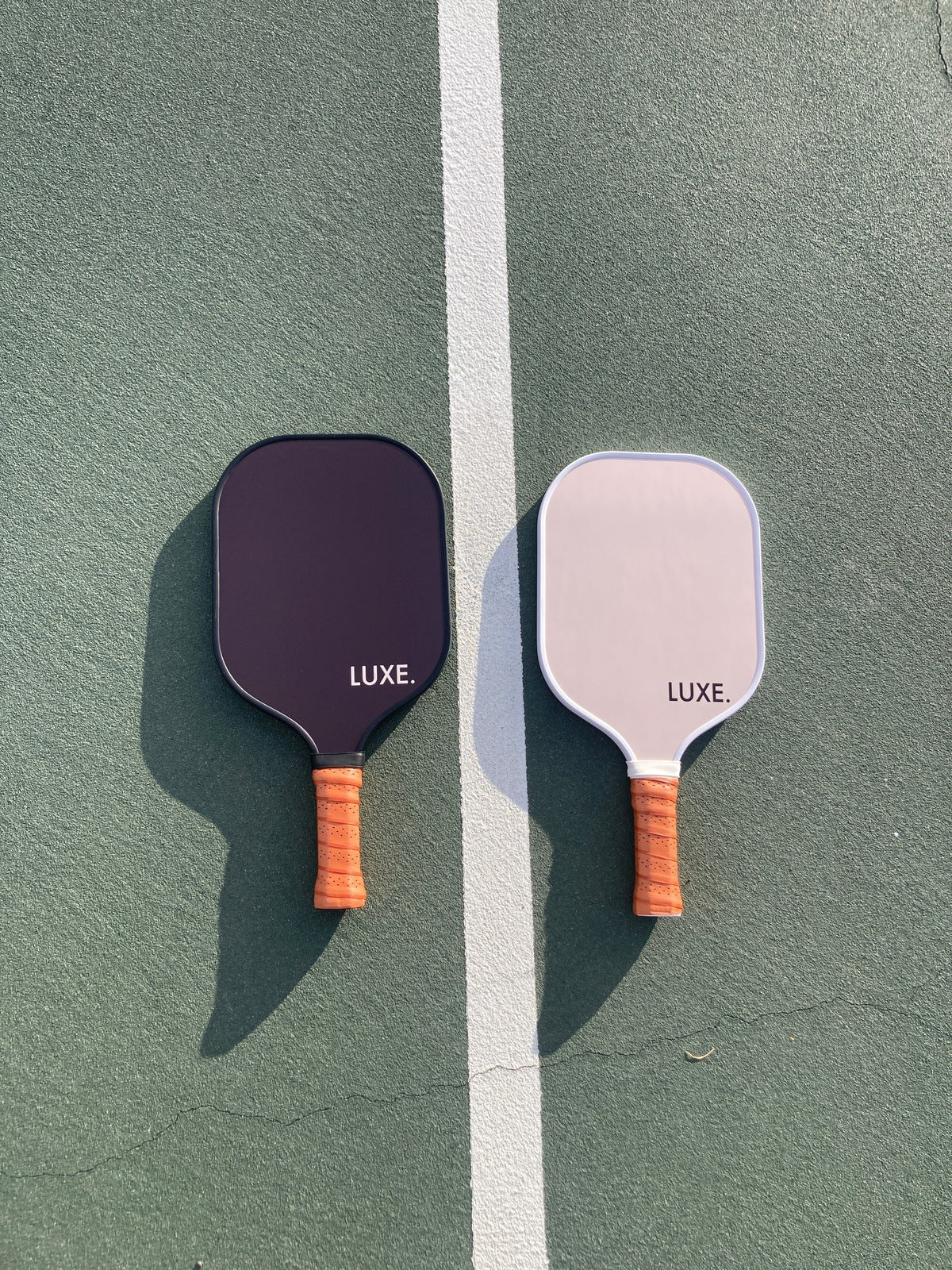 Two pickleball paddles on a green court with a white line separating them. The paddle on the left is black with a brown handle, and the one on the right is light pink with a brown handle. Both have the word "LUXE." printed on them.