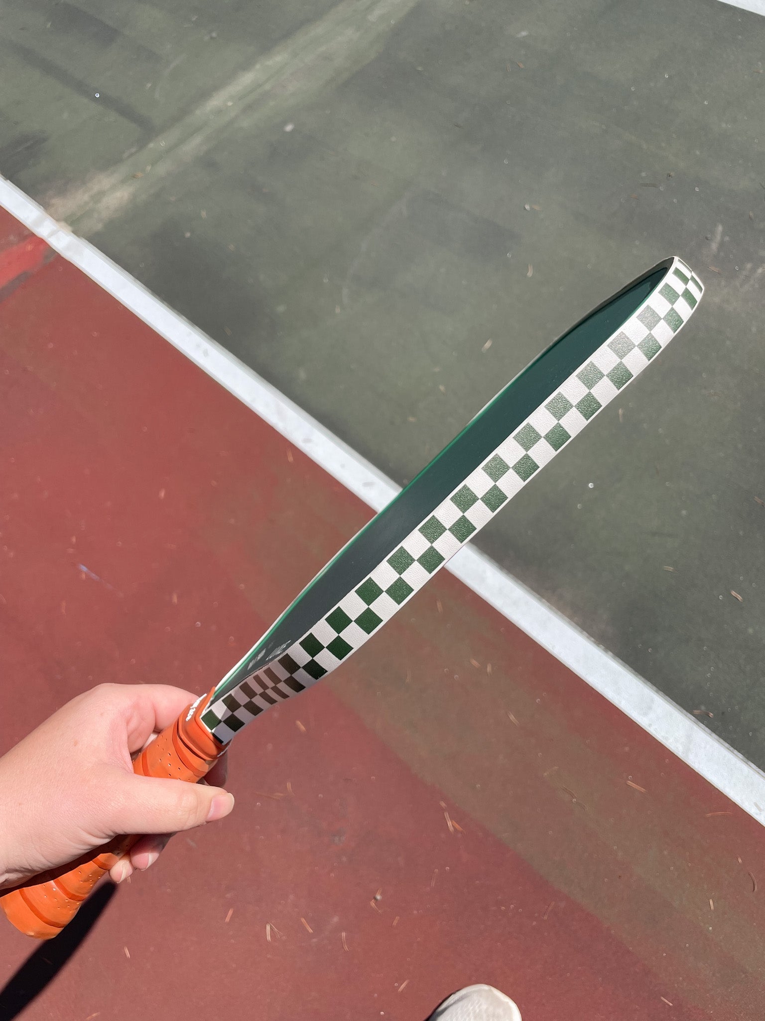 Hand holding a green and white checkered pickleball paddle with an orange grip on a tennis court.
