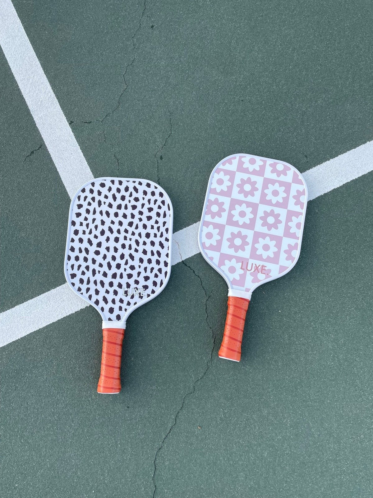 Two pickleball paddles, one with black spots and one with pink flowers, lie on a green pickleball court.