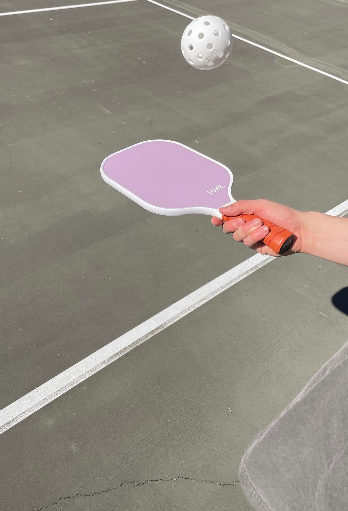 A person holding a lavender pickleball paddle with a white ball in the air on a green pickleball court.