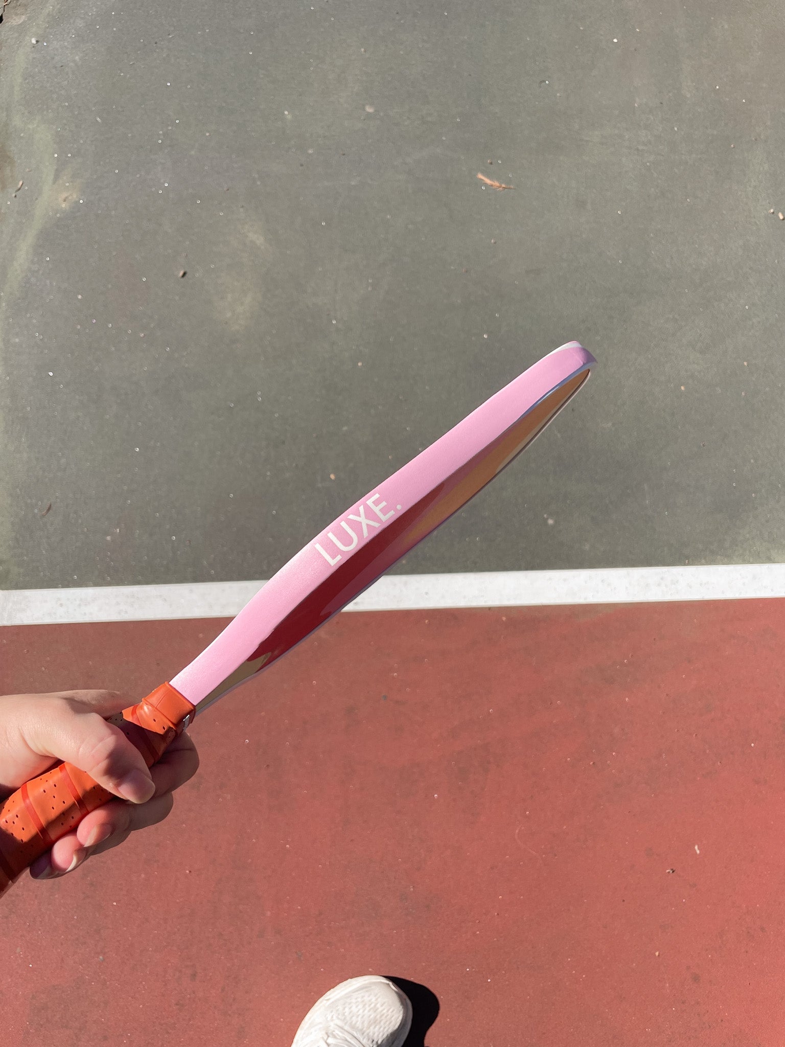 Close-up of a hand holding a pink Luxe pickleball paddle on a red and gray court.