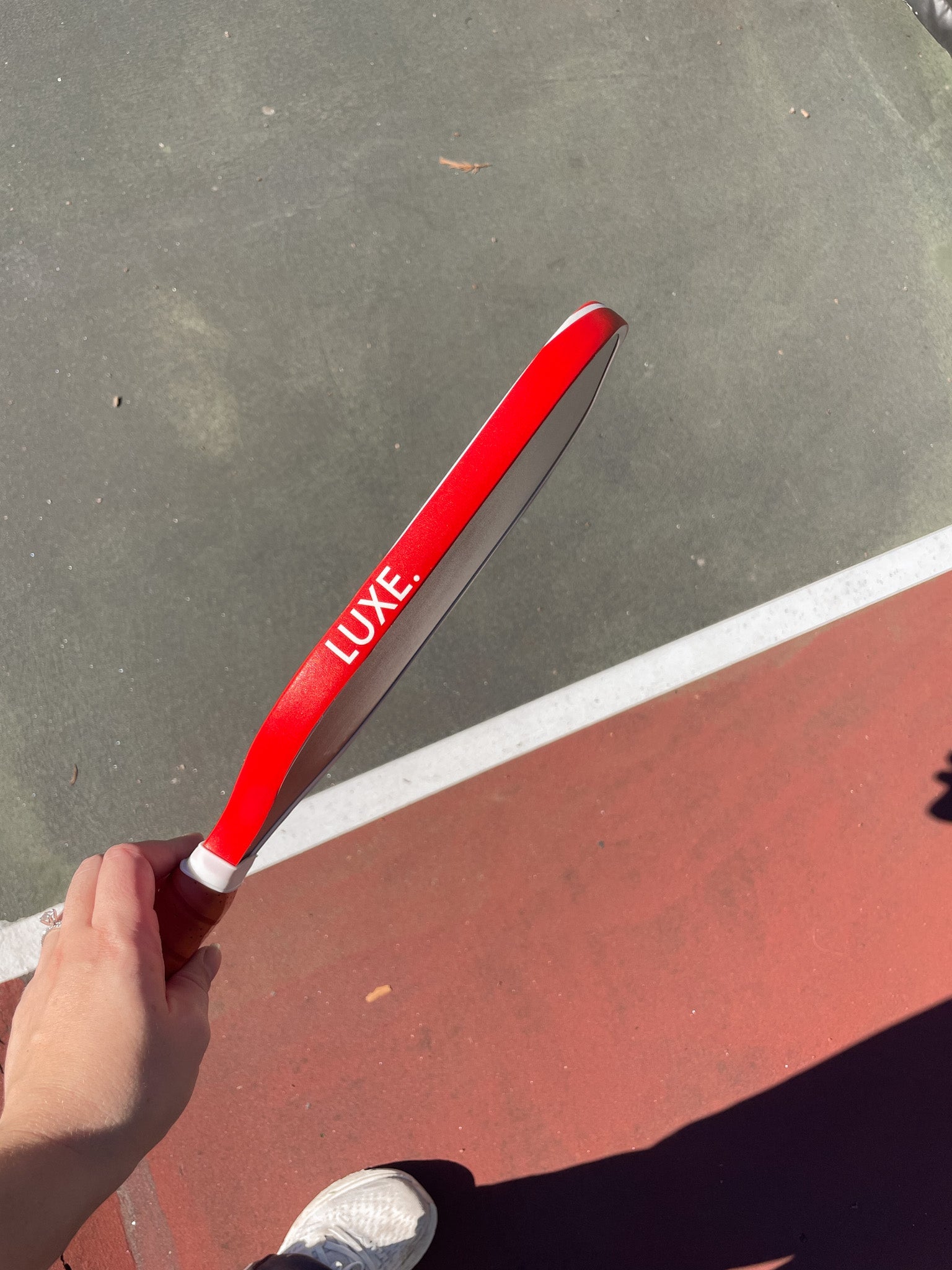 Hand holding a red "LUXE" pickleball paddle on a red and gray court.