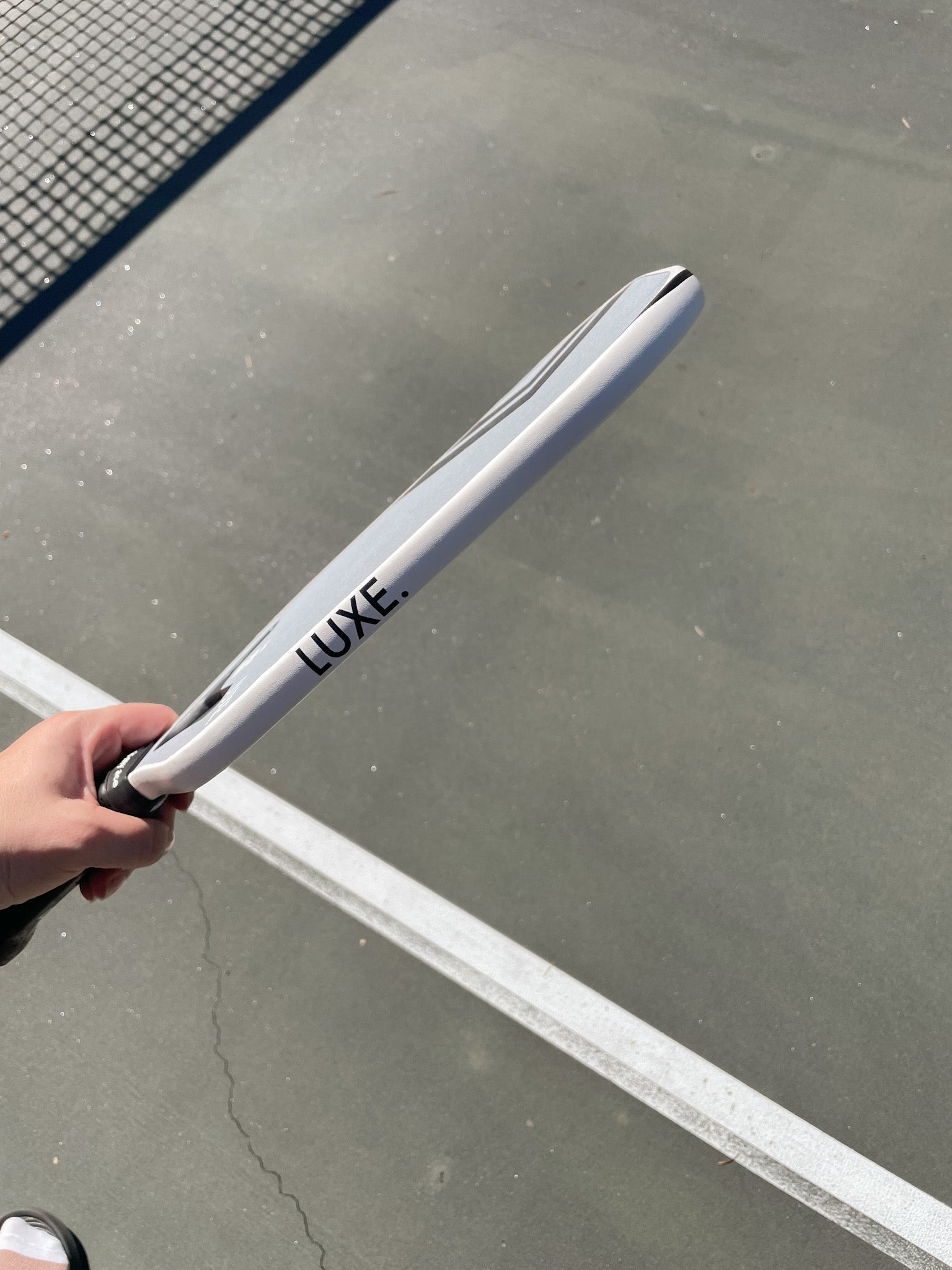 A hand holds a LUXE pickleball paddle on an outdoor court.
