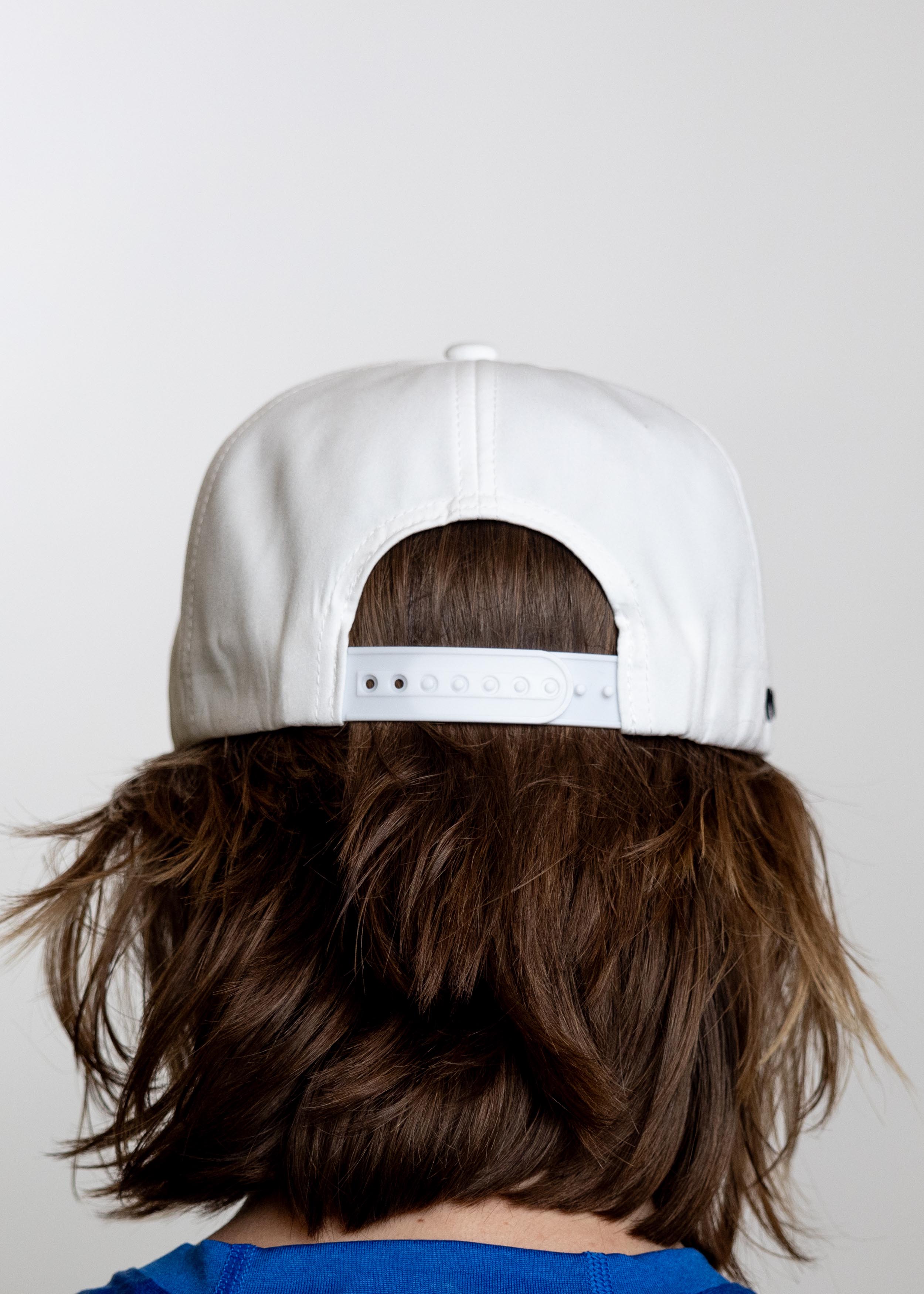 White flat bill cap by TMPR Sports, back view, with adjustable snapback closure, on brown hair above a blue shirt.