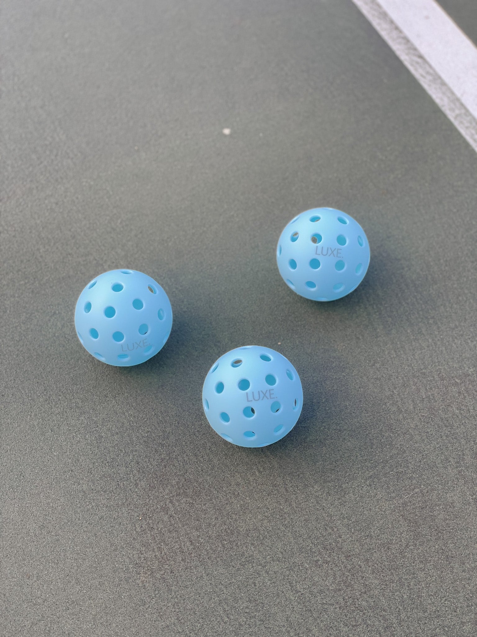 Three light blue pickleballs with "LUXE" branding on a grey court.