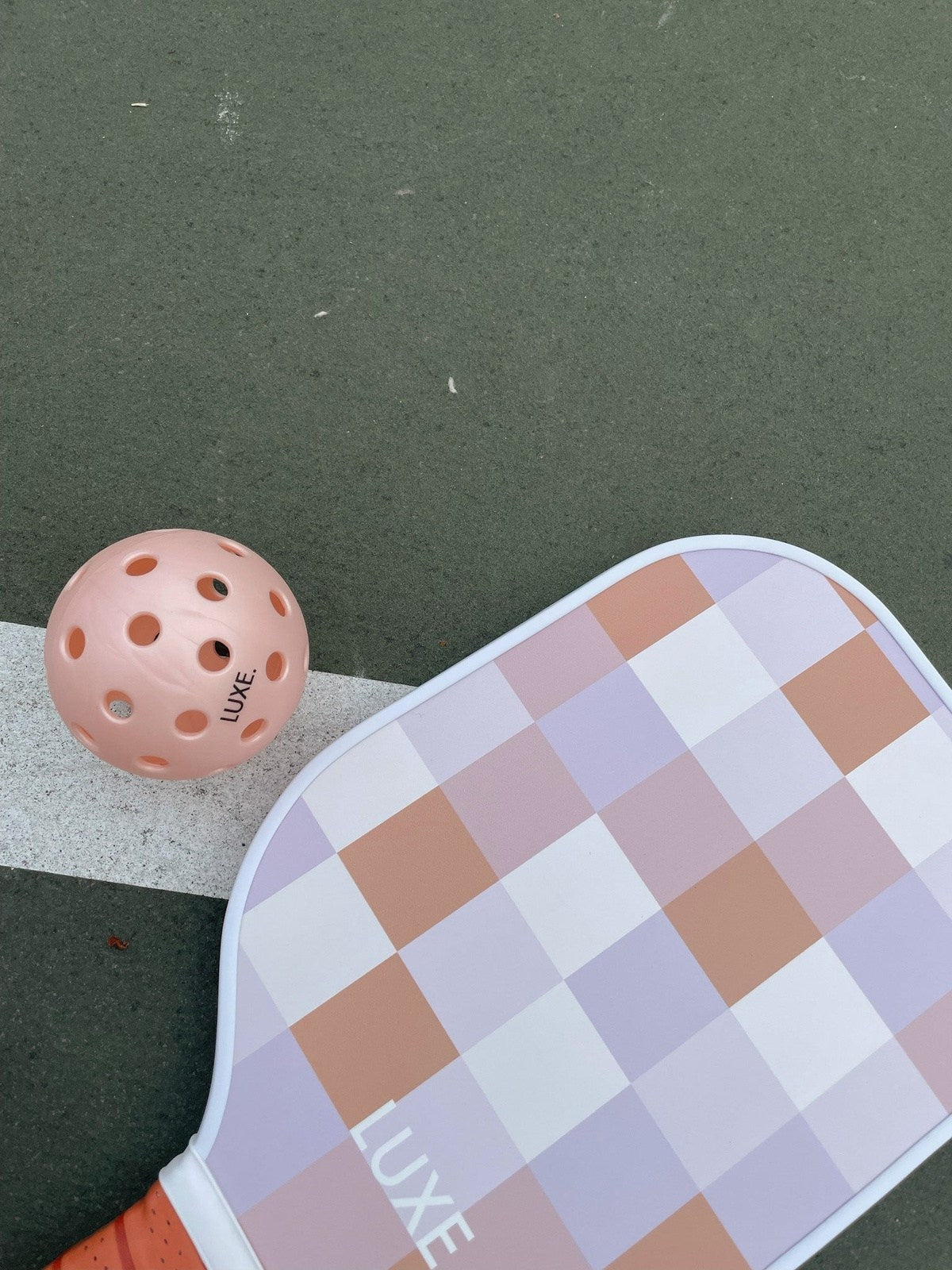 A pink pickleball and a checkered pickleball paddle rest on a green and white court.