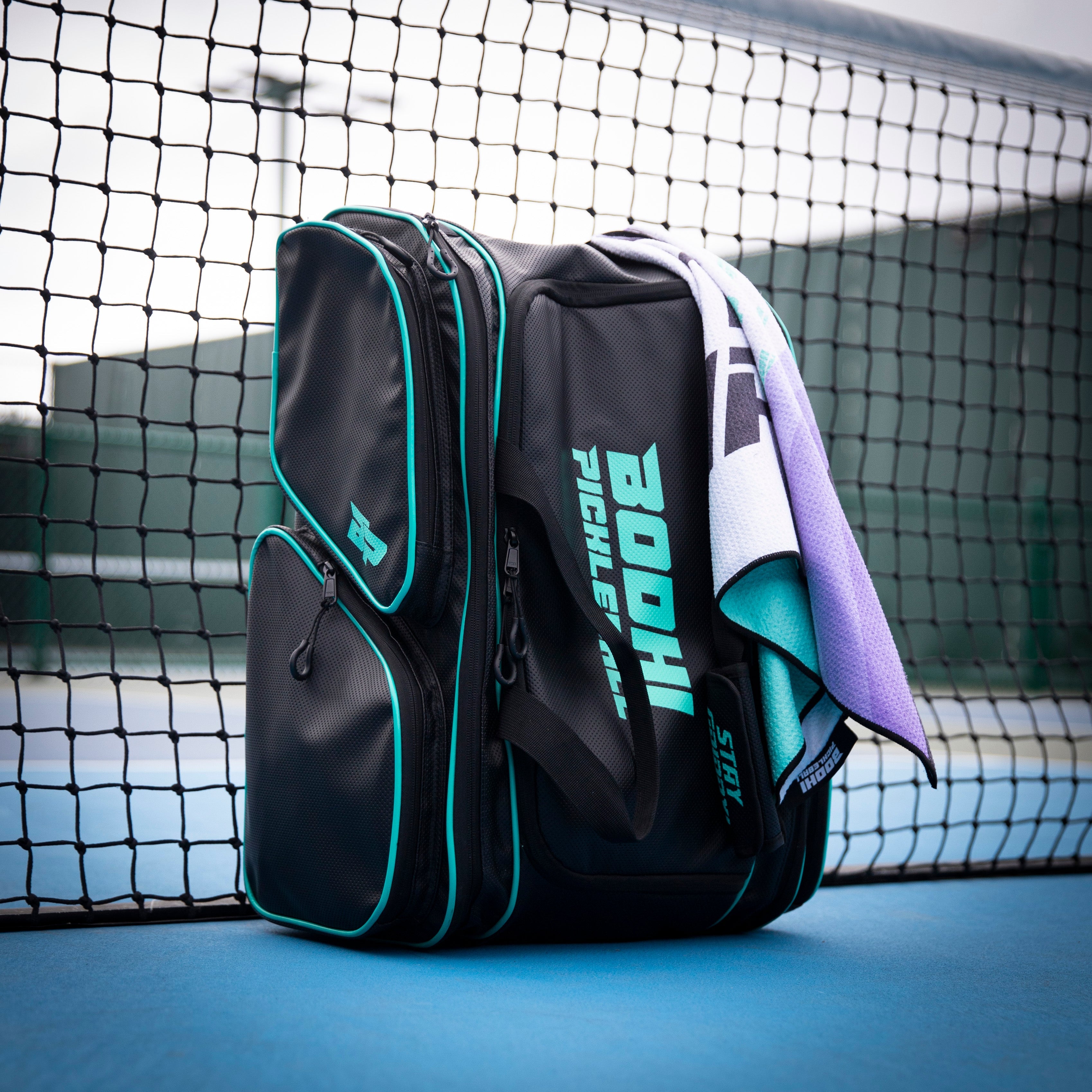 Black pickleball bag with teal trim and a towel, on a blue court by a net.