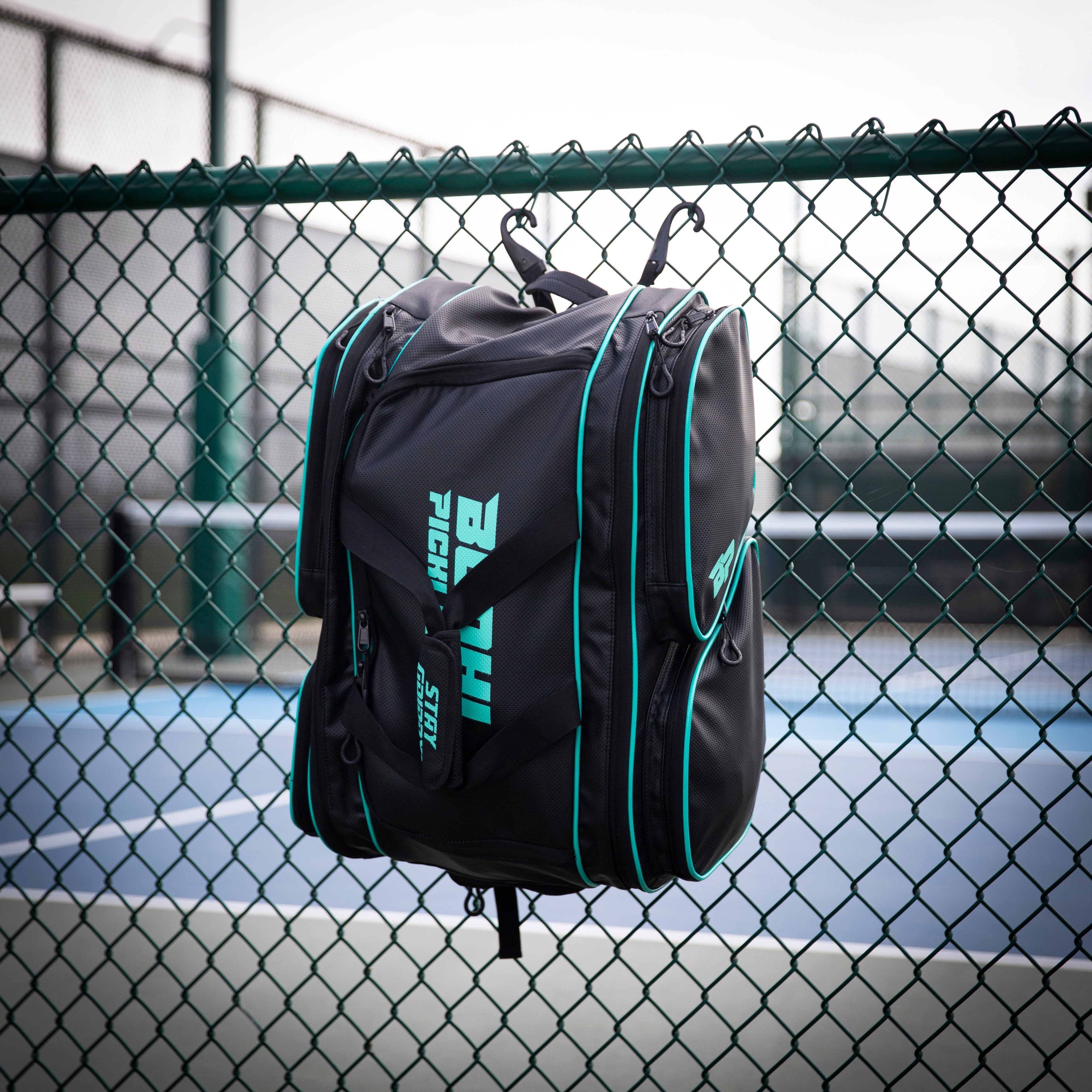 Black tennis bag with teal accents hangs on a green chain-link fence.