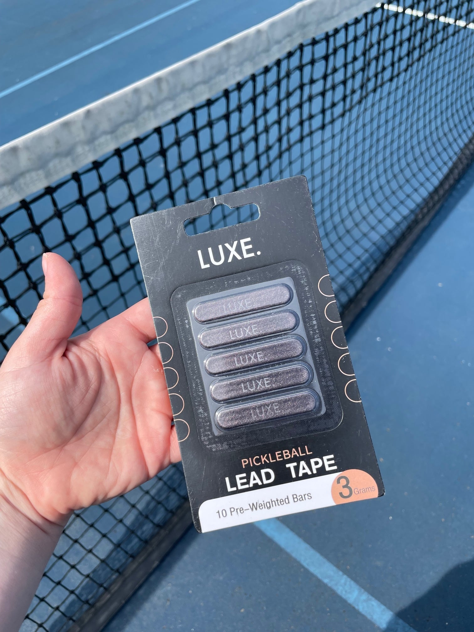 A hand holds a package of Luxe Pickleball Lead Tape with 10 pre-weighted 3-gram bars against a blue tennis or pickleball court with a net in the background.