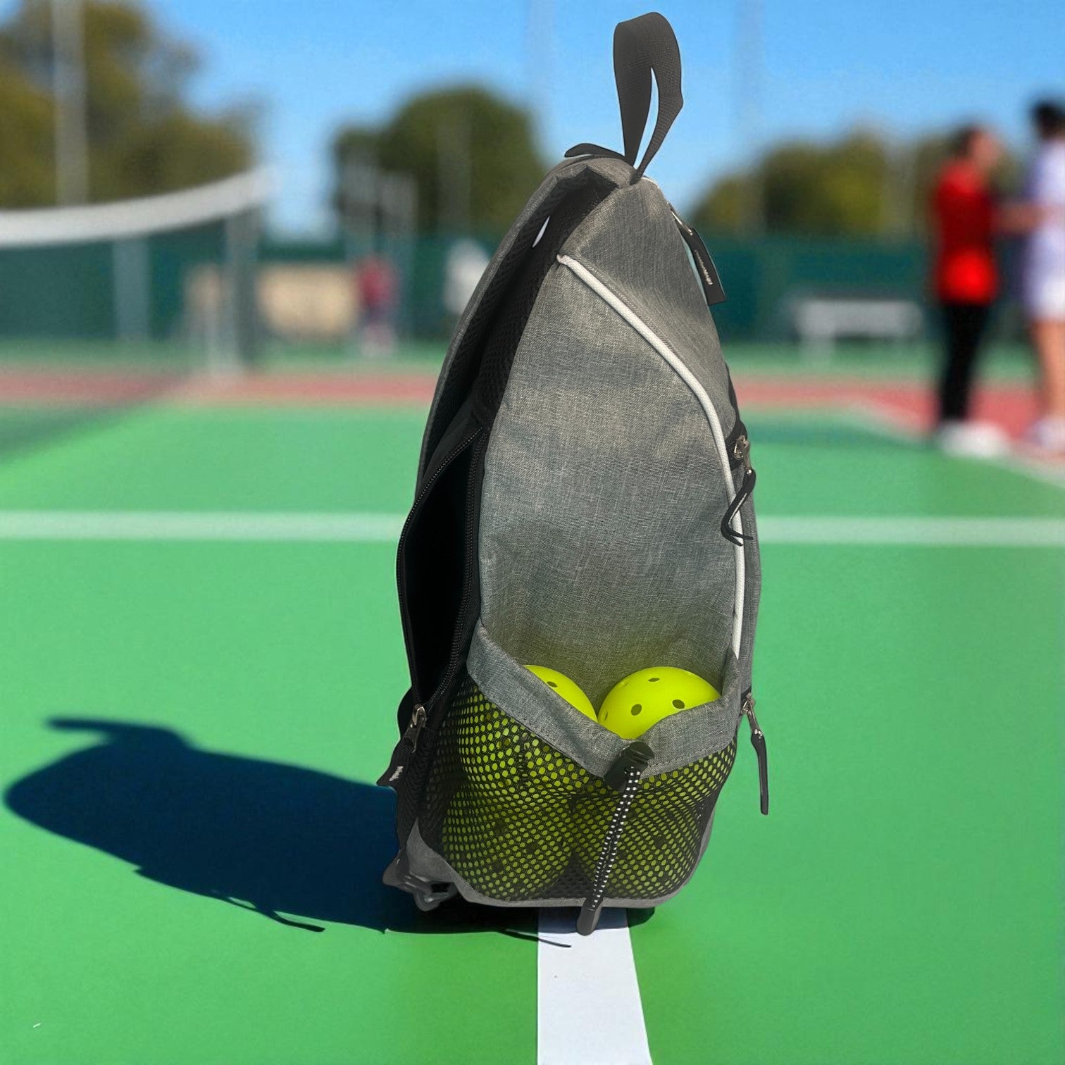 Gray pickleball backpack with two yellow pickleballs in the mesh pocket. The backpack is on a green court.