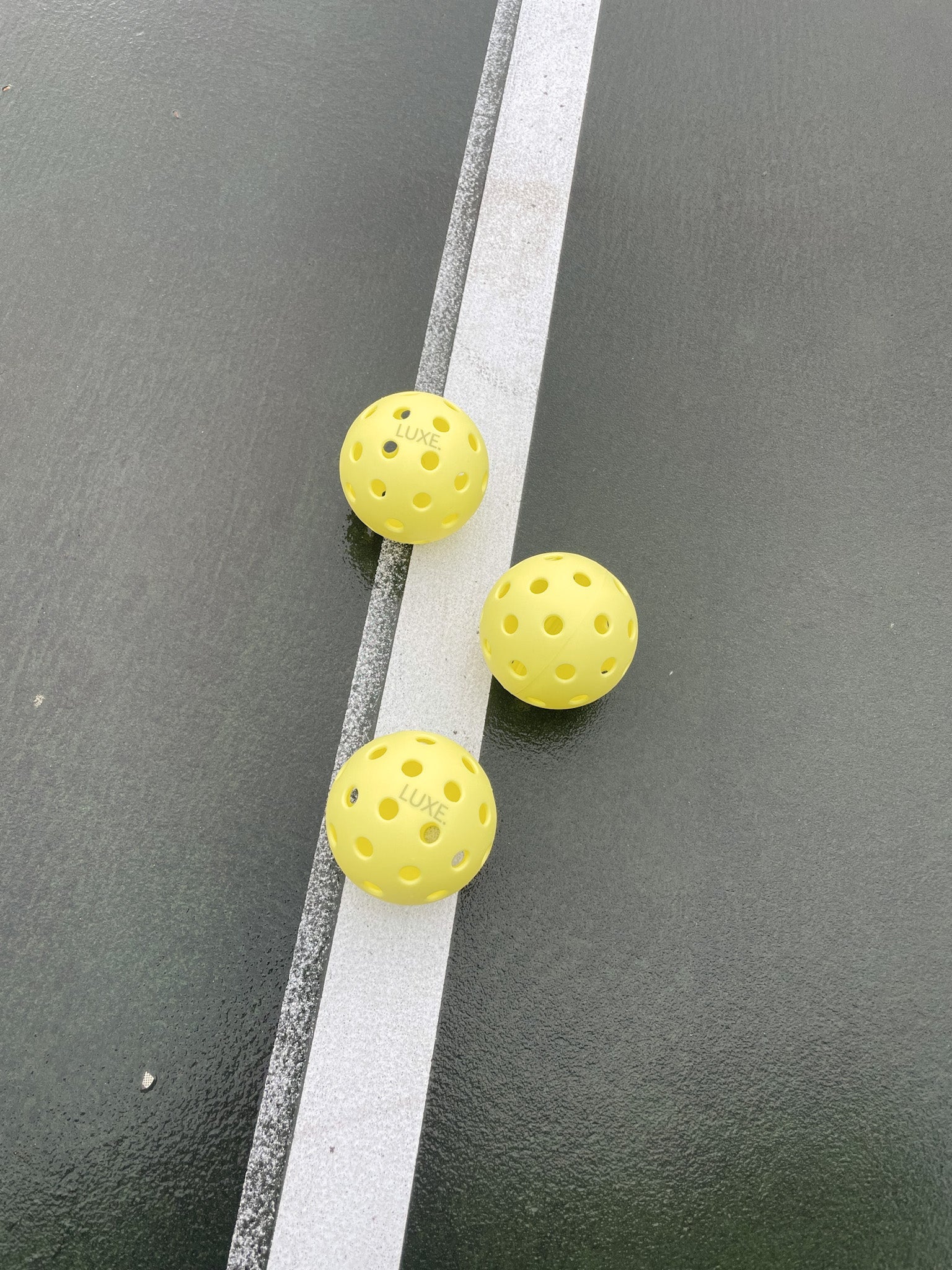 Three yellow pickleballs with "LUXE" printed on them, resting on a gray surface intersected by a white line.