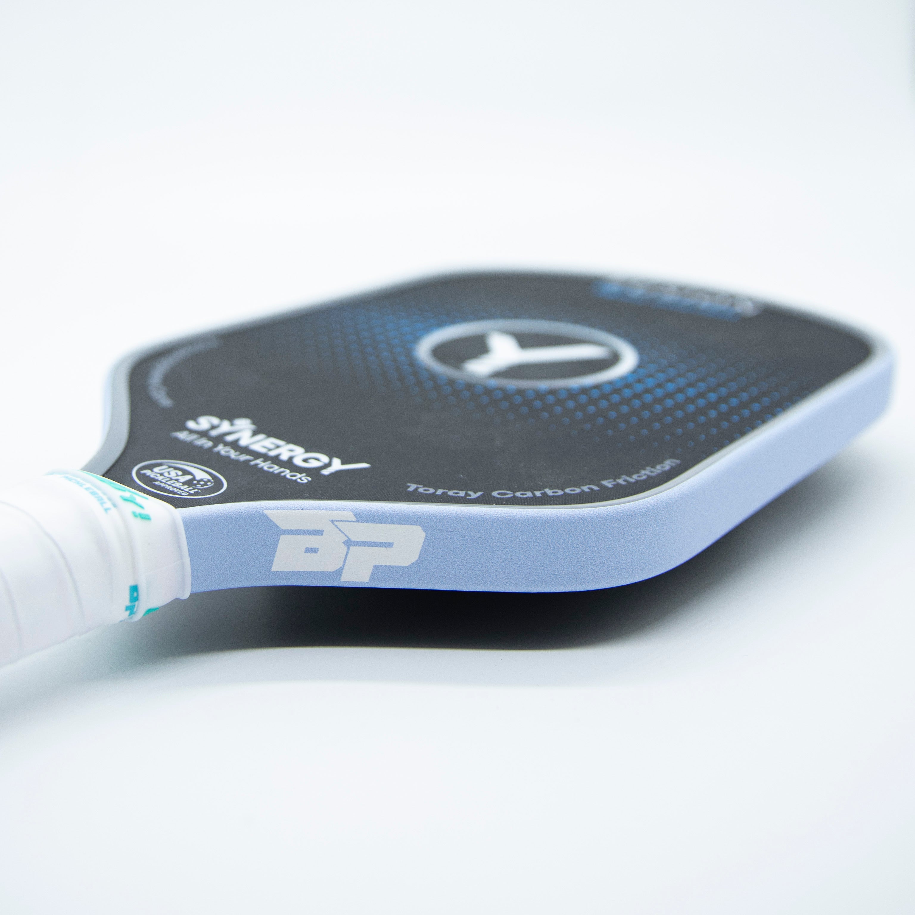 Angled black pickleball paddle by DinkersAlley.com, with a light blue edge guard, white grip, 'SYNERGY' text, and BP logo.