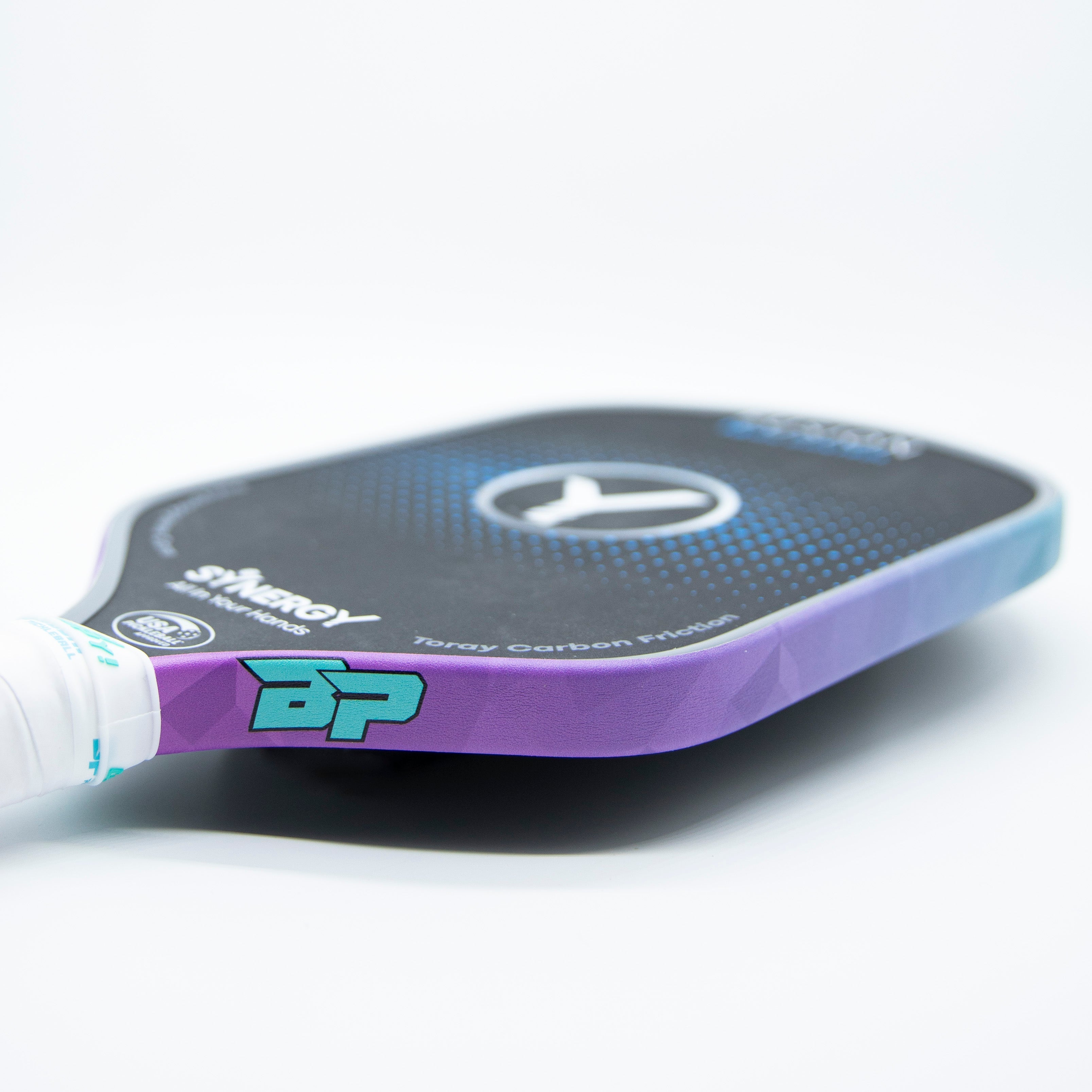 A close-up of a pickleball paddle with a white grip, a purple and teal handle with a "BP" logo, and a black paddle face with a blue dot pattern and a white "Y" logo.