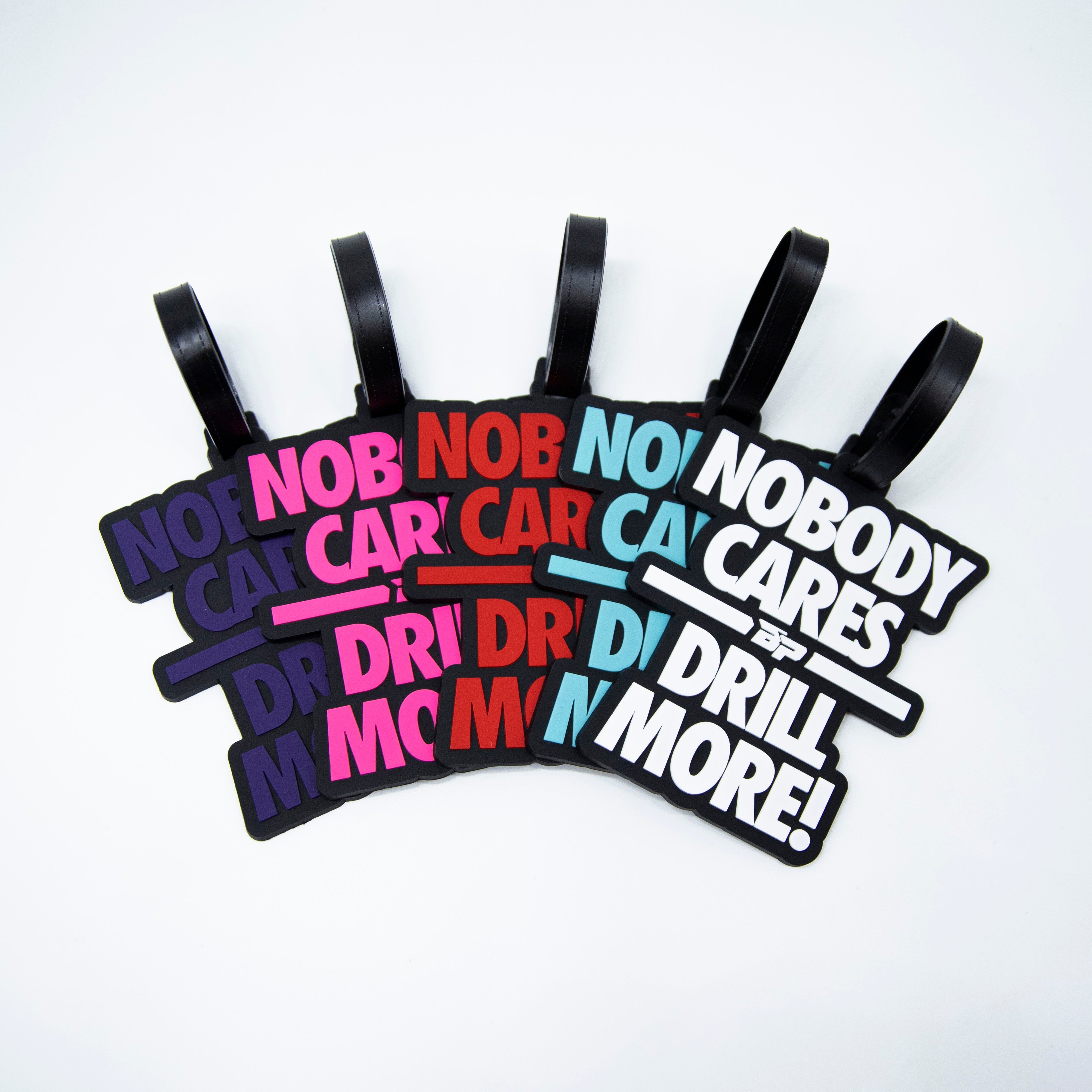 Five colorful luggage tags with black straps reading "Nobody cares, drill more!" on a white background.