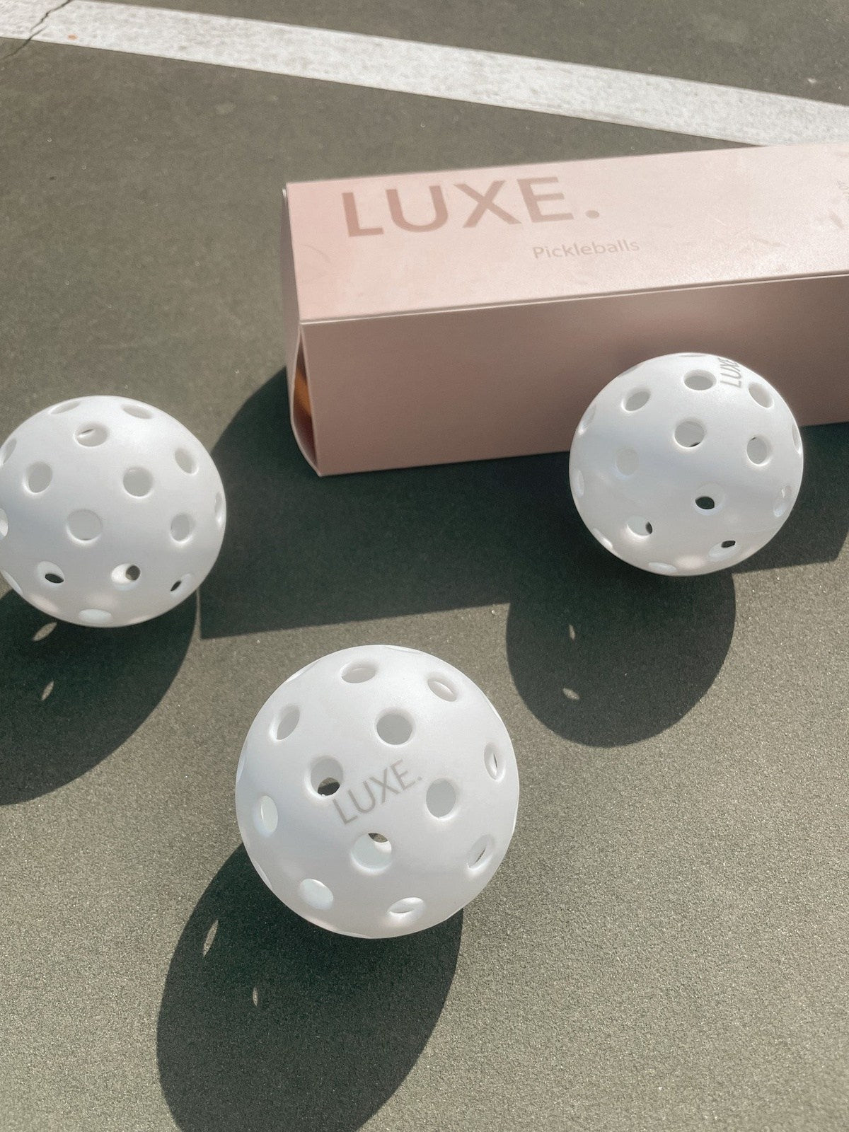Three white pickleballs and a pink Luxe pickleball box on a pickleball court.