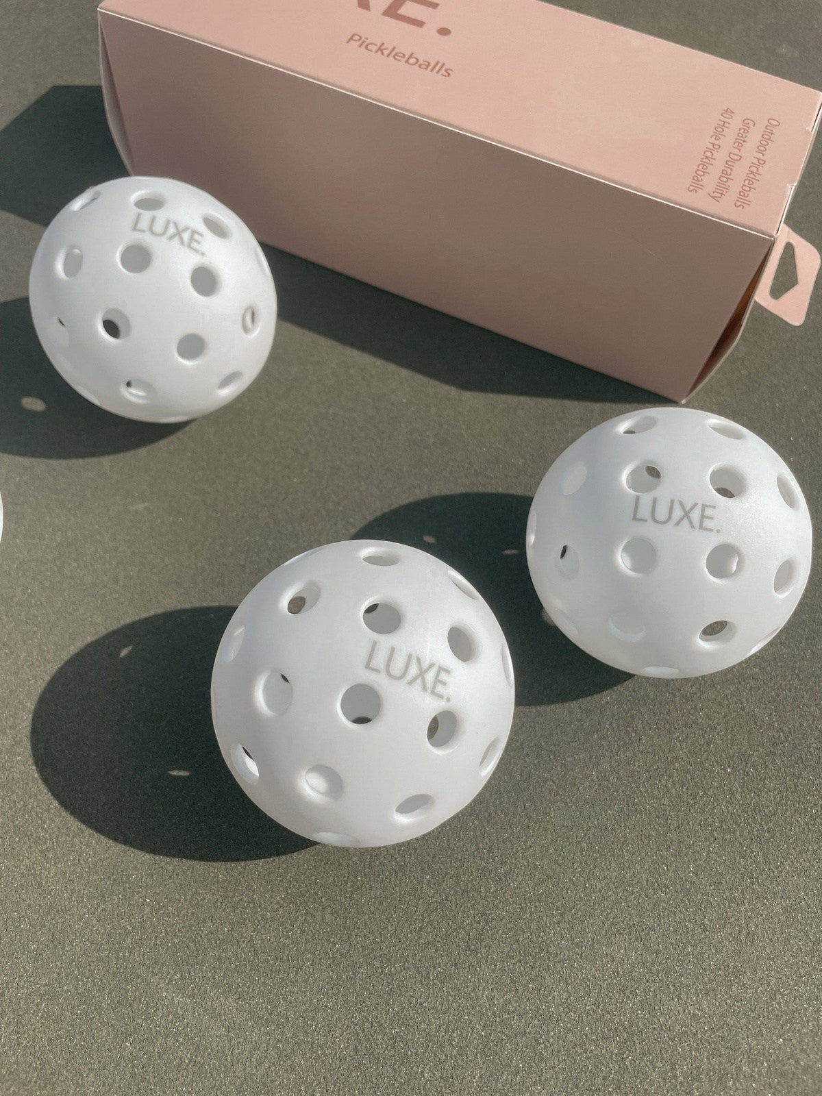 Three white pickleballs with the word "LUXE" next to a pink box.